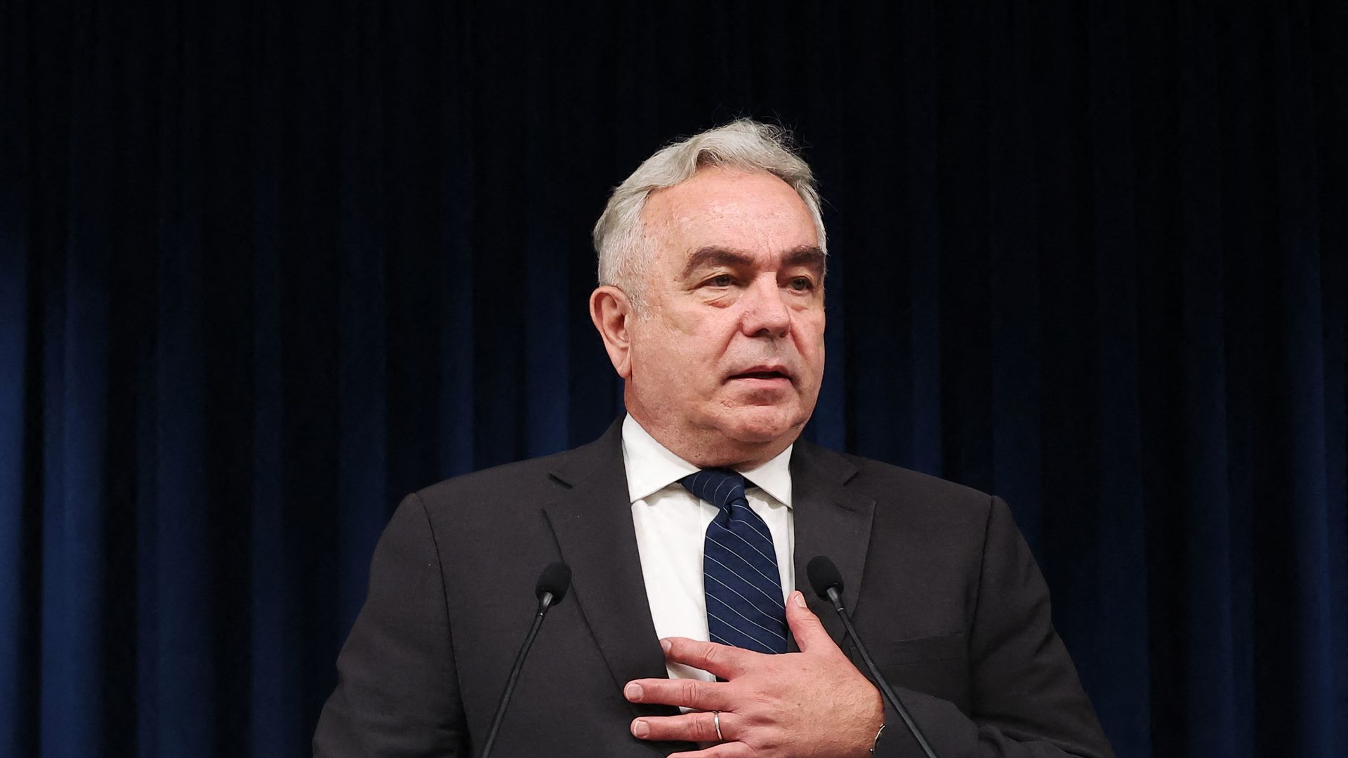 A white-haired man in a grey suit holds is left hand to his chest in a gesture of empathy