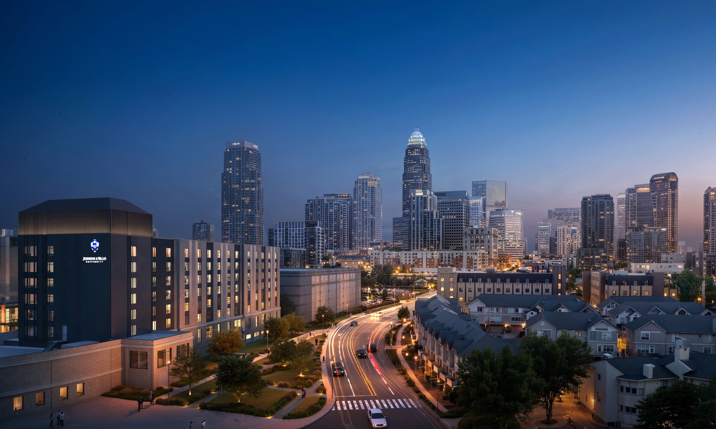 City skyline at dusk with tall glass towers; blue gradient sky. Left foreground shows Johnson & Wales University building; curved highway with light trails winds past low-rise homes.