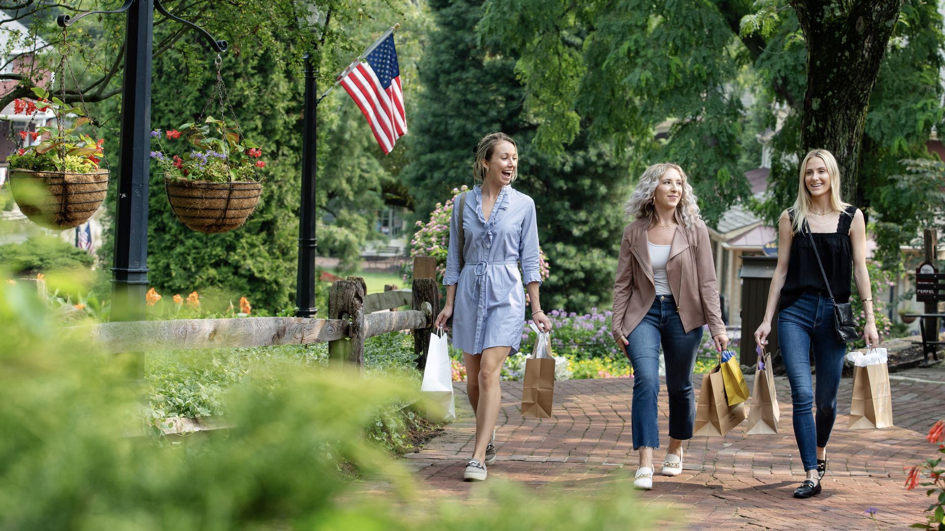 Three women stroll along a brick garden path, smiling and carrying shopping bags. They pass hanging flower baskets, a lamppost with an American flag, and dense green trees.