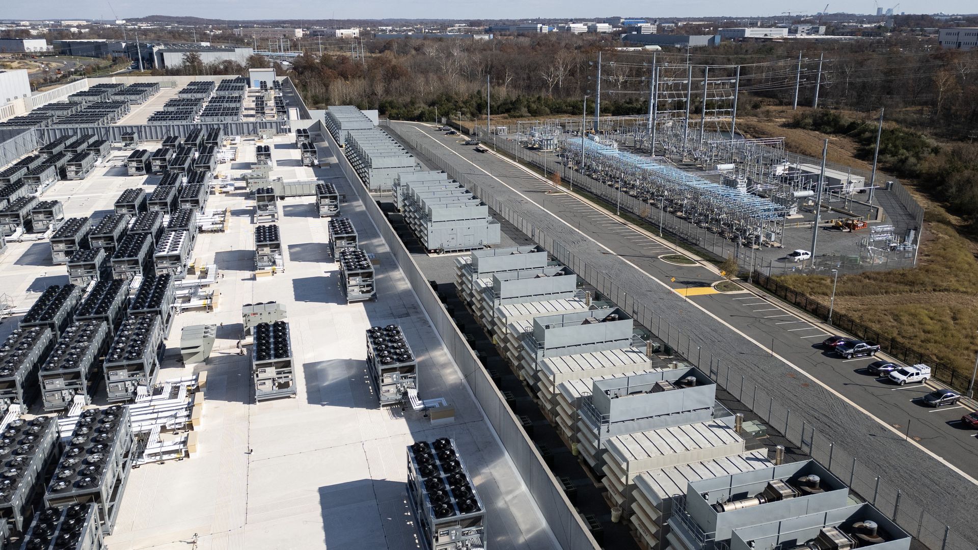 Aerial view of a large data center roof with rows of cooling units and fans; to the right is a fenced substation with transformers, a road with parked cars, and a distant cityscape under a blue sky.