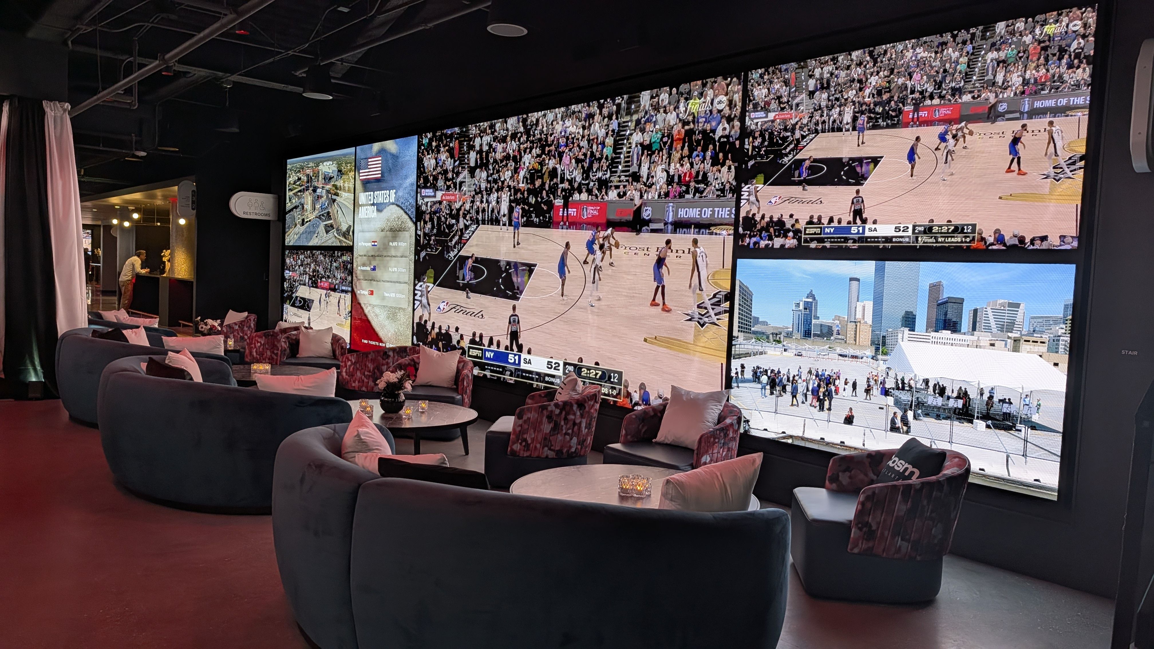 A lounge with a curved wall of large video screens showing a crowded basketball game and arena scoreboard; plush gray couches with pink cushions, round coffee tables, and dim lighting.