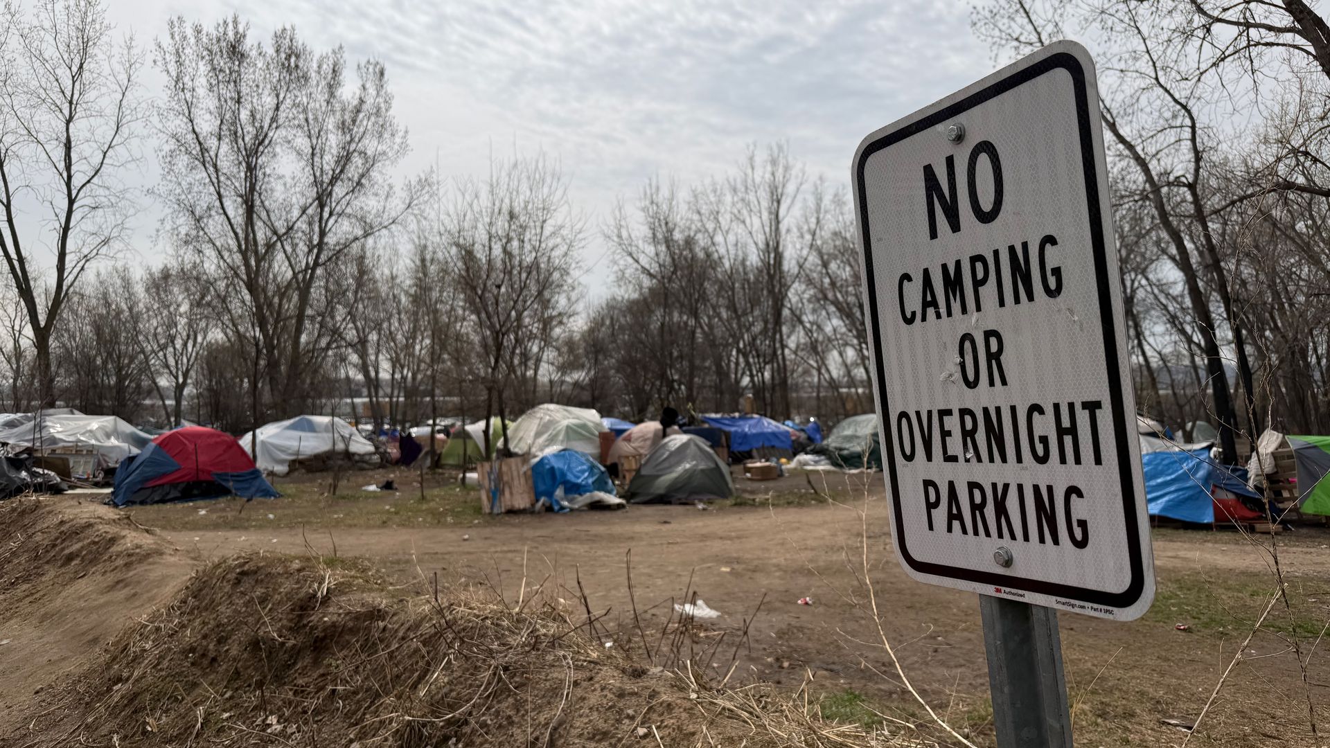 Wide outdoor scene with a large sign in the foreground reading "NO CAMPING OR OVERNIGHT PARKING"; behind it, a cluttered encampment of tents among bare trees under a cloudy sky.