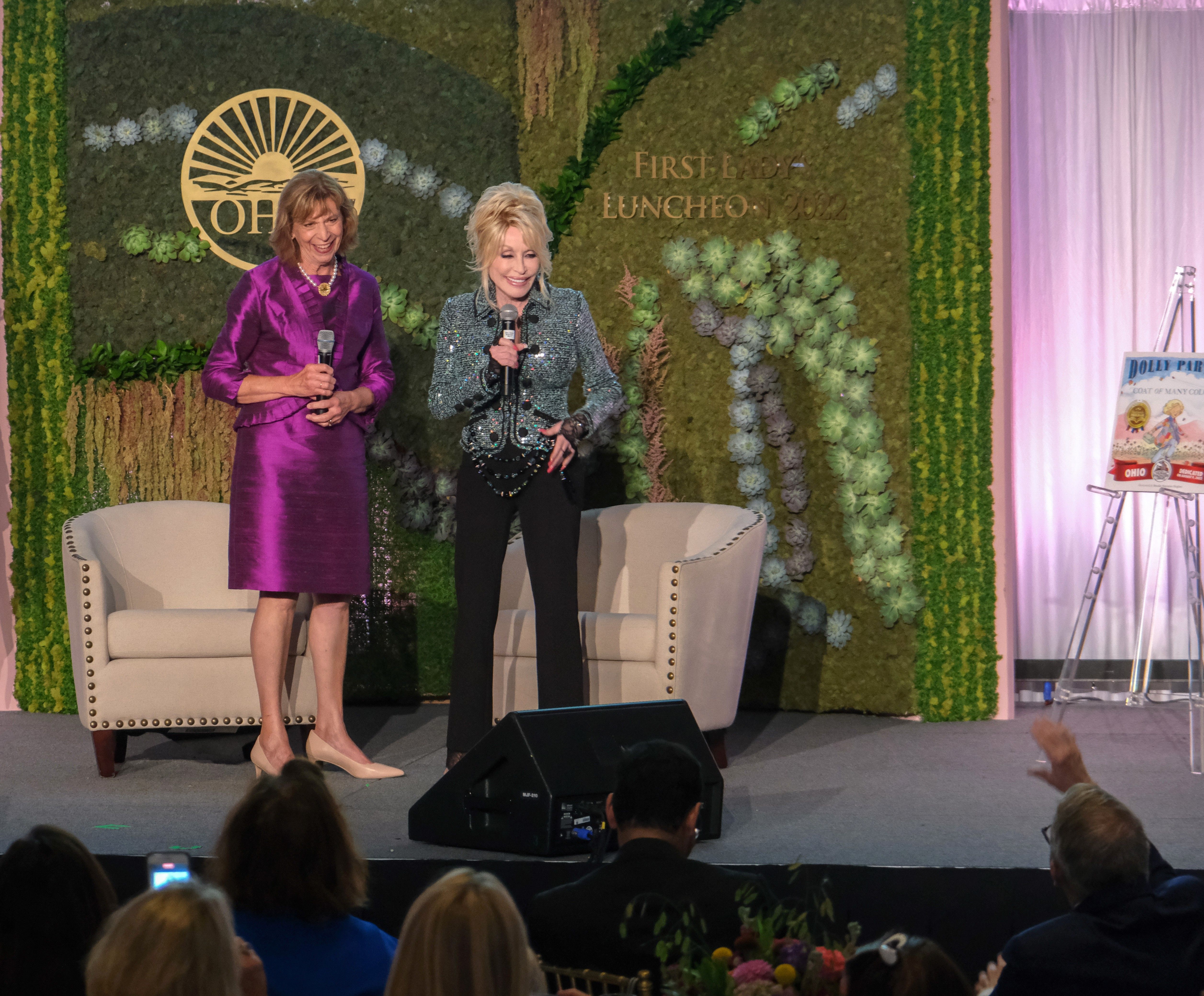 Ohio First Lady Fran DeWine stands next to Dolly Parton on a stage with two chairs and a sign that reads "First Lady Luncheon" in the background. 