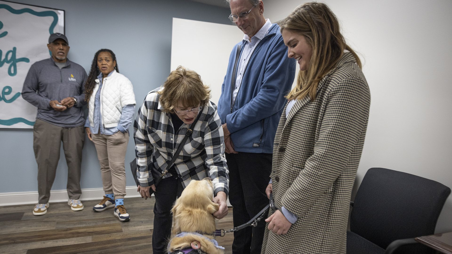 Five people in a room interact with a golden service dog wearing a purple vest. One woman bends down to pet the dog, others smile or watch. Casual clothing, wood floor.