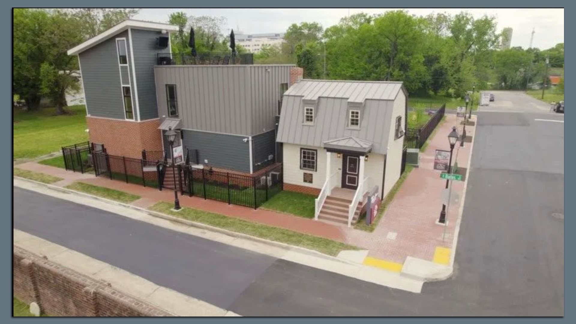 Aerial view of a modern two-building complex on a street corner: a tall gray-brick structure with a black fence on the left, connected to a white-and-gray cottage with a metal roof on the right; trees in background.