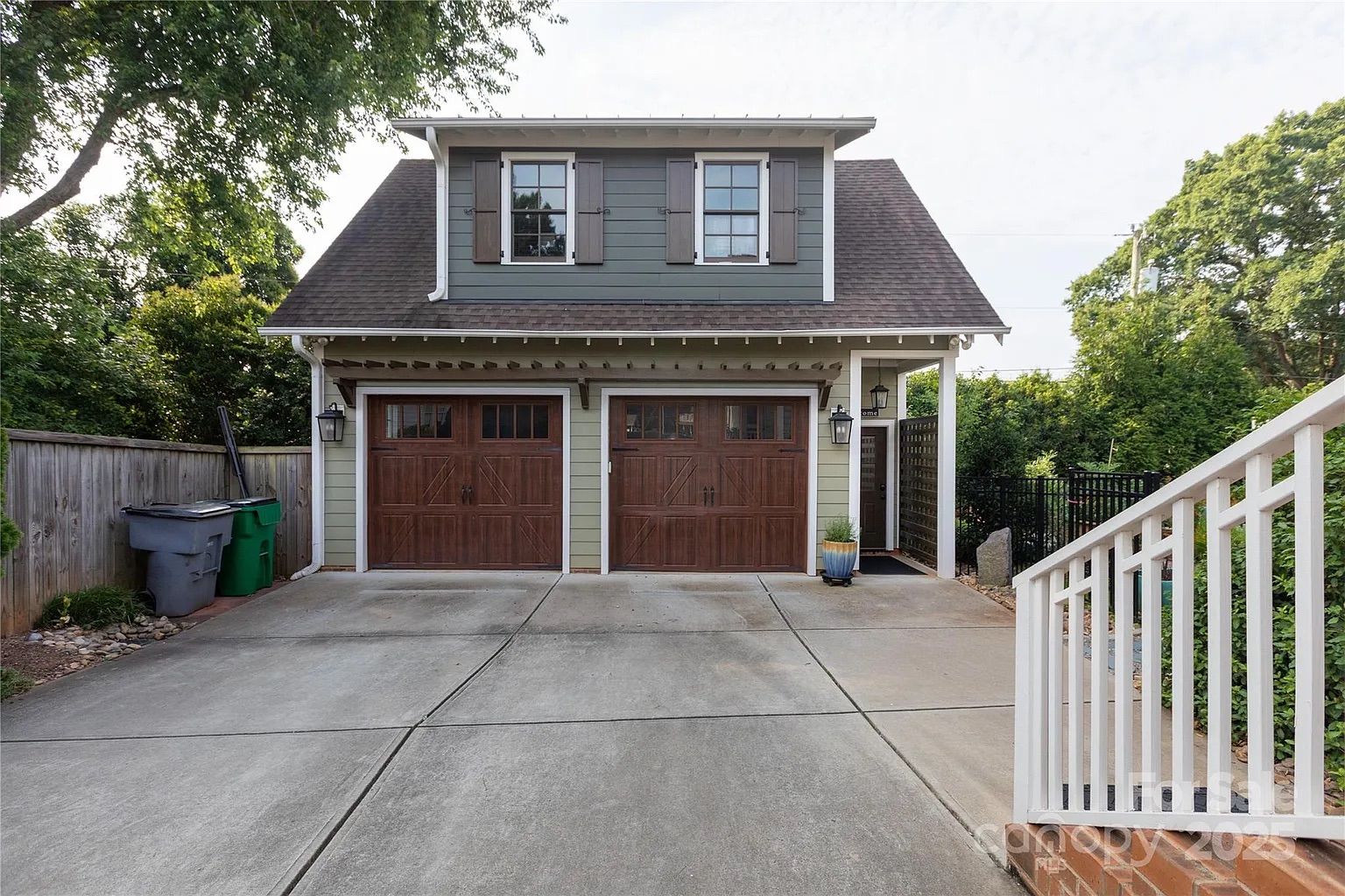 Two-story garage with gray siding, brown shingled roof, and two wooden garage doors. Concrete driveway in front, trash bins on the left, and white railing on the right with green trees in background.
