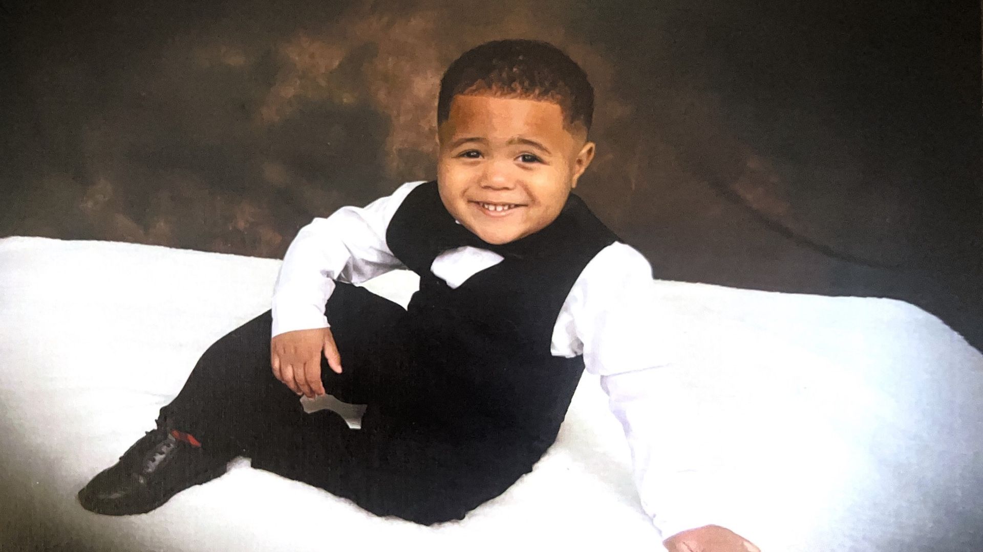 A posed photo of Reese Jackson smiling at the camera while reclined on white furniture wearing formalwear.
