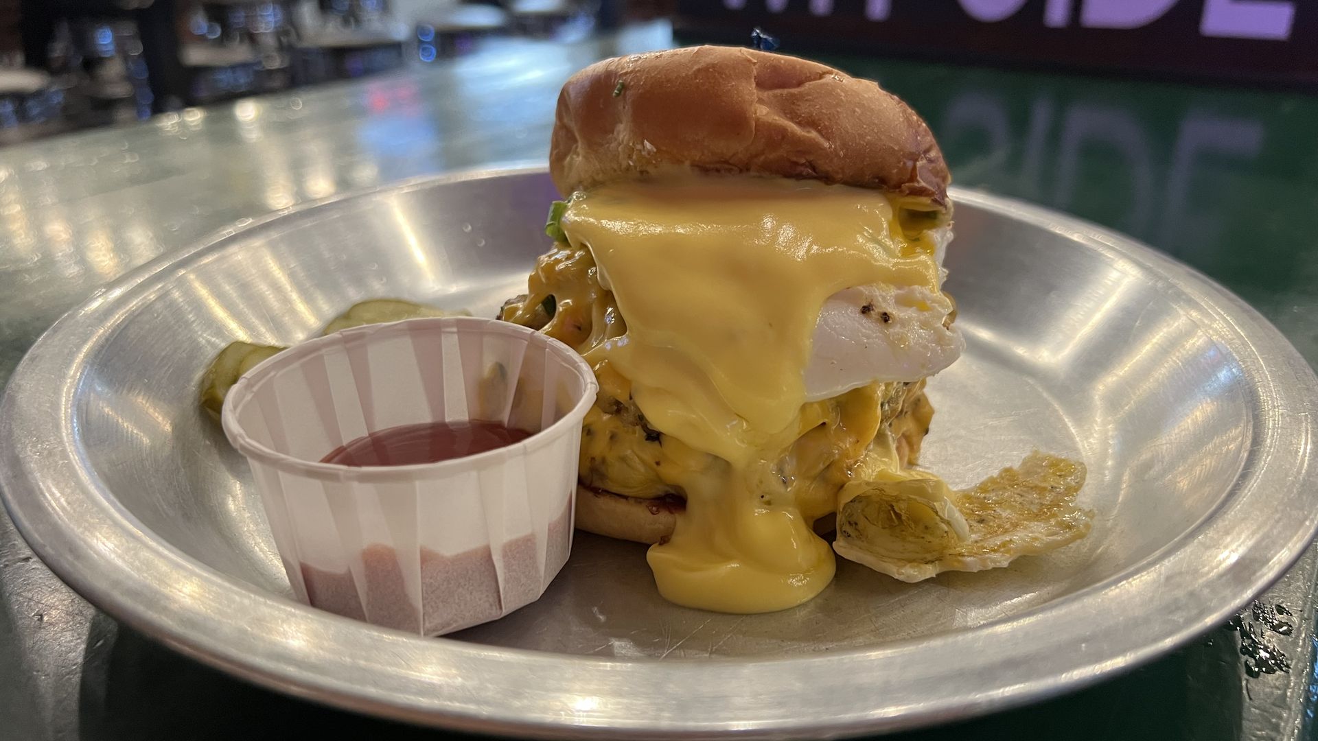 Cheeseburger with hollandaise sauce and a fried egg on a metal plate, served with pickles and a small cup of ketchup on the side