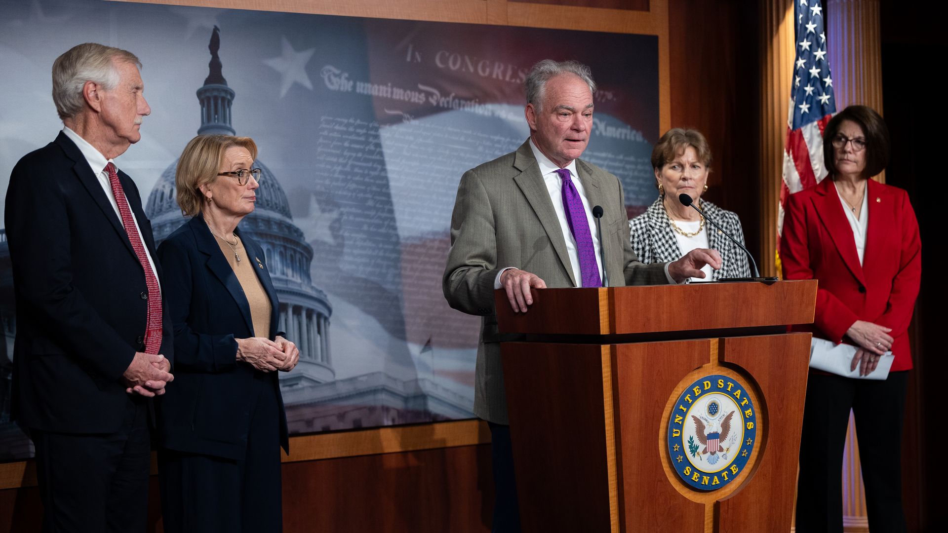 Tim Kaine, wearing a gray suit and a purple tie, speaks at a podium while flanked by several other lawmakers.