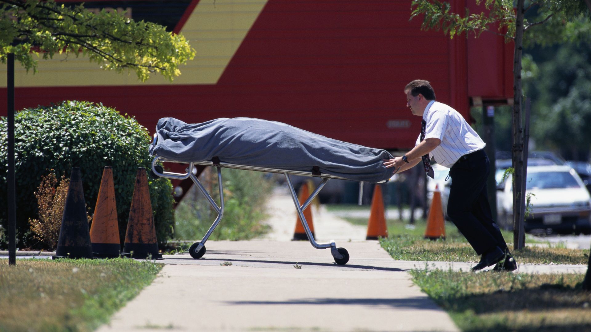 A man in a white shirt pushing a gurnee with a corpse on it.