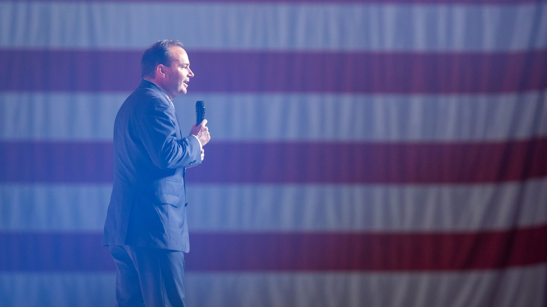 Sen. Mike Lee speaking on the microphone with an American flag backdrop. 