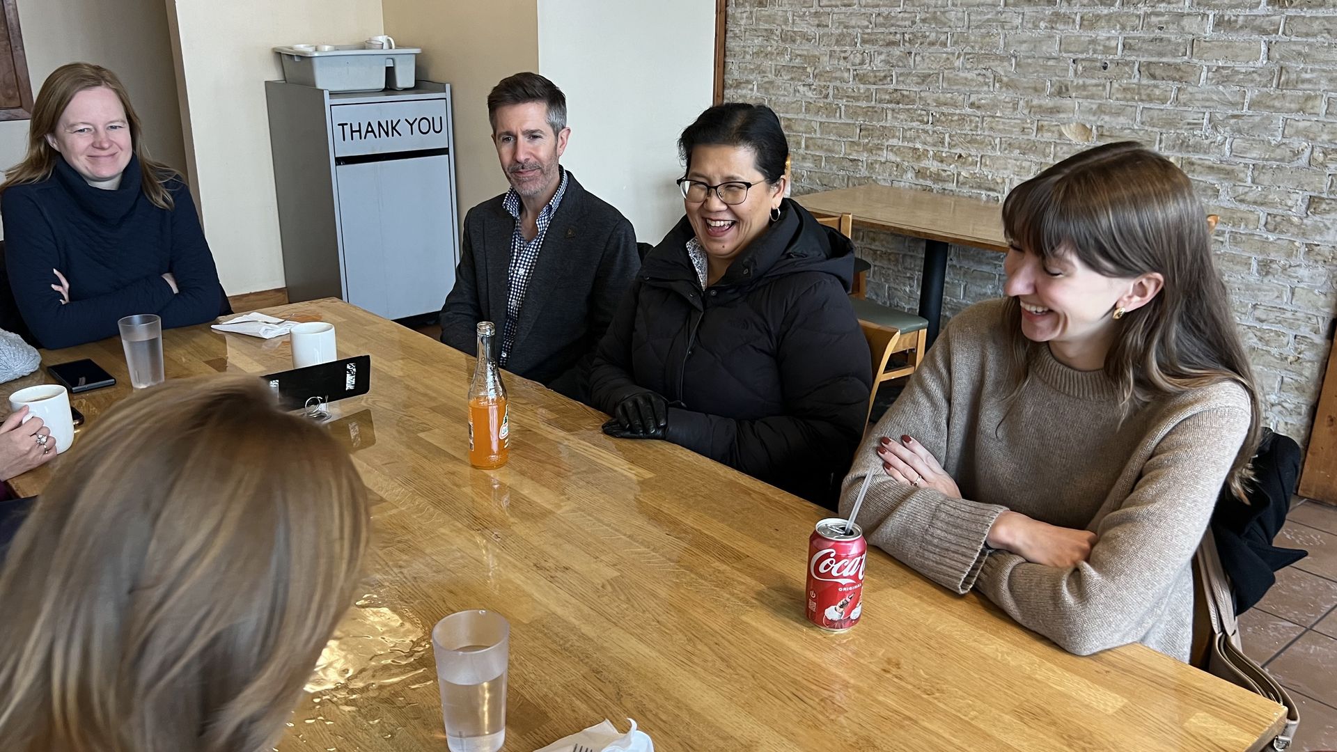Five people sitting around a wooden table in a casual dining setting, smiling and engaged in conversation, with drinks including a Coca-Cola can and an orange soda bottle on the table.