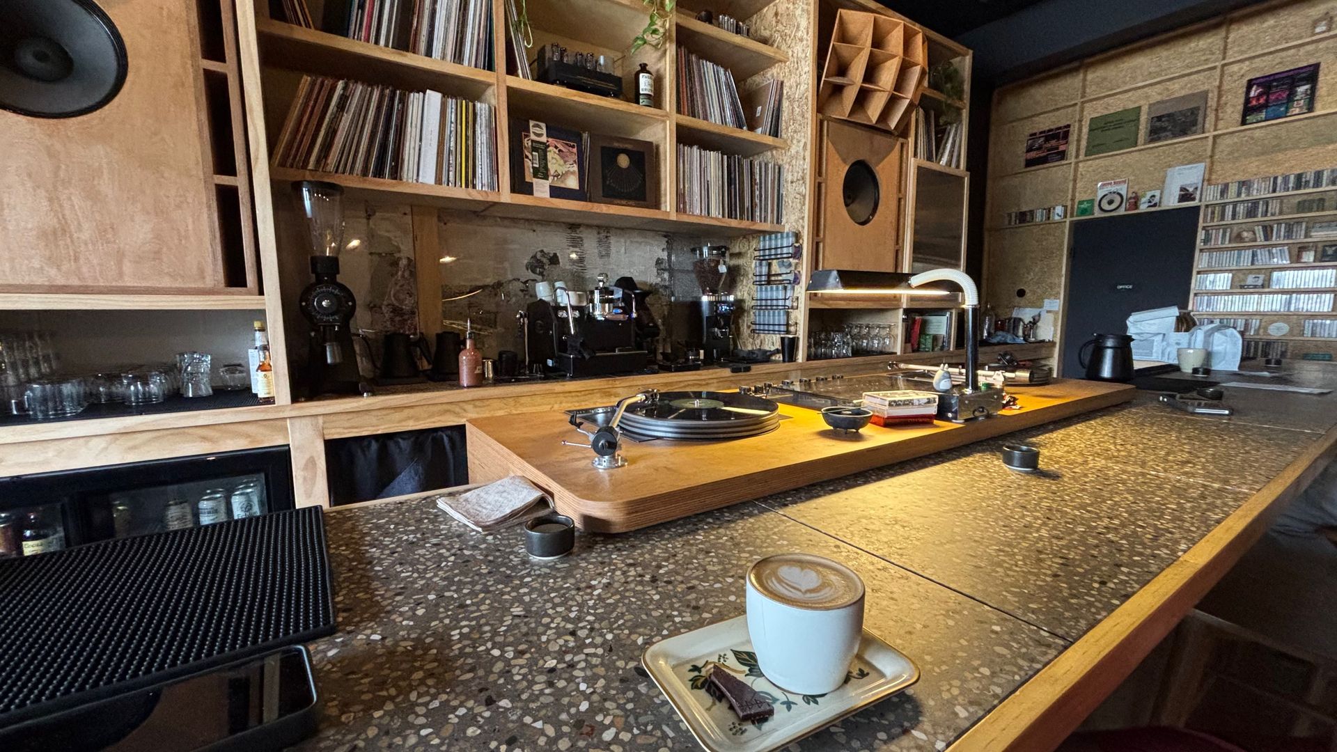 Cozy retro bar with a terrazzo counter, vinyl records on wooden shelves, espresso setup, and a turntable on a wooden board. A latte in a white cup on a floral saucer sits foreground.
