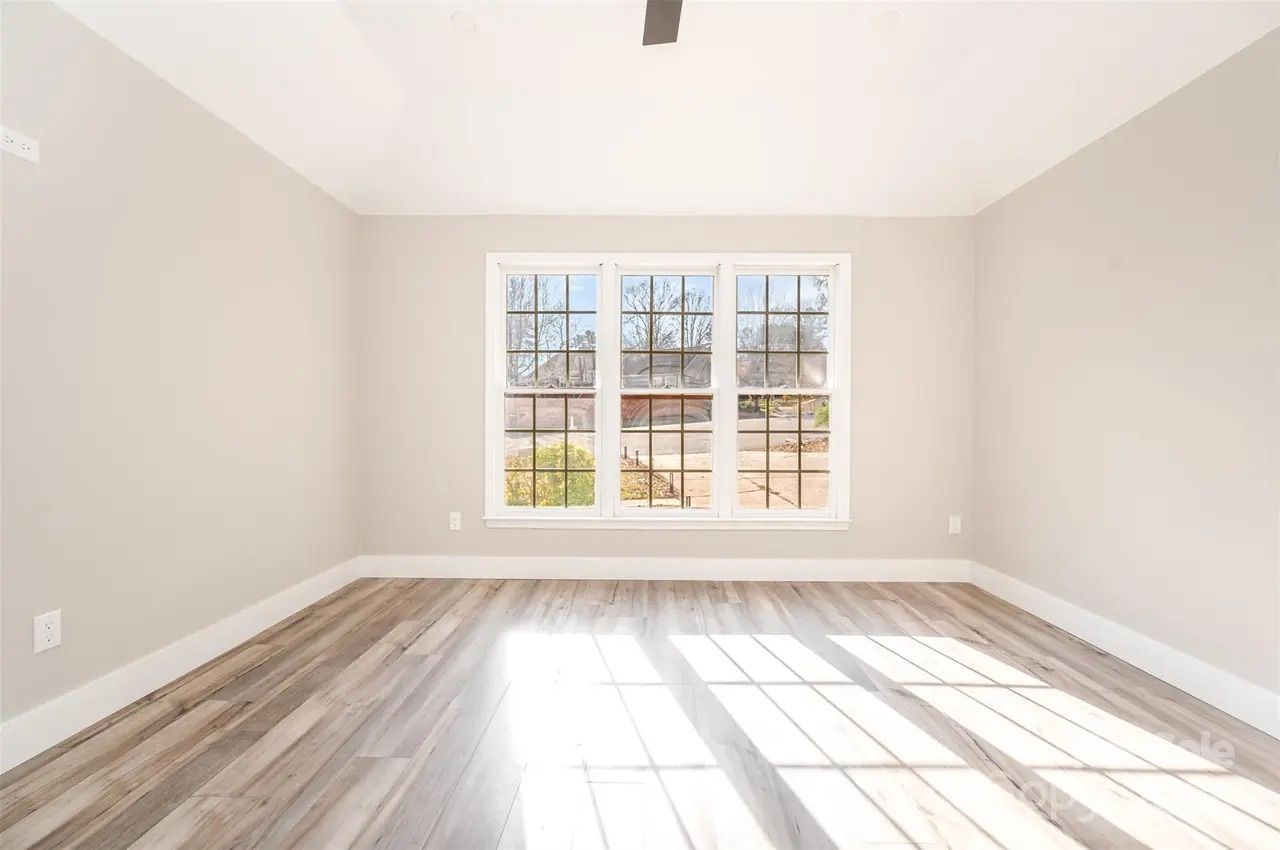 Empty room with light wood flooring, beige walls, white trim, a large three-section window letting in bright sunlight and casting shadows on the floor, and a ceiling fan.