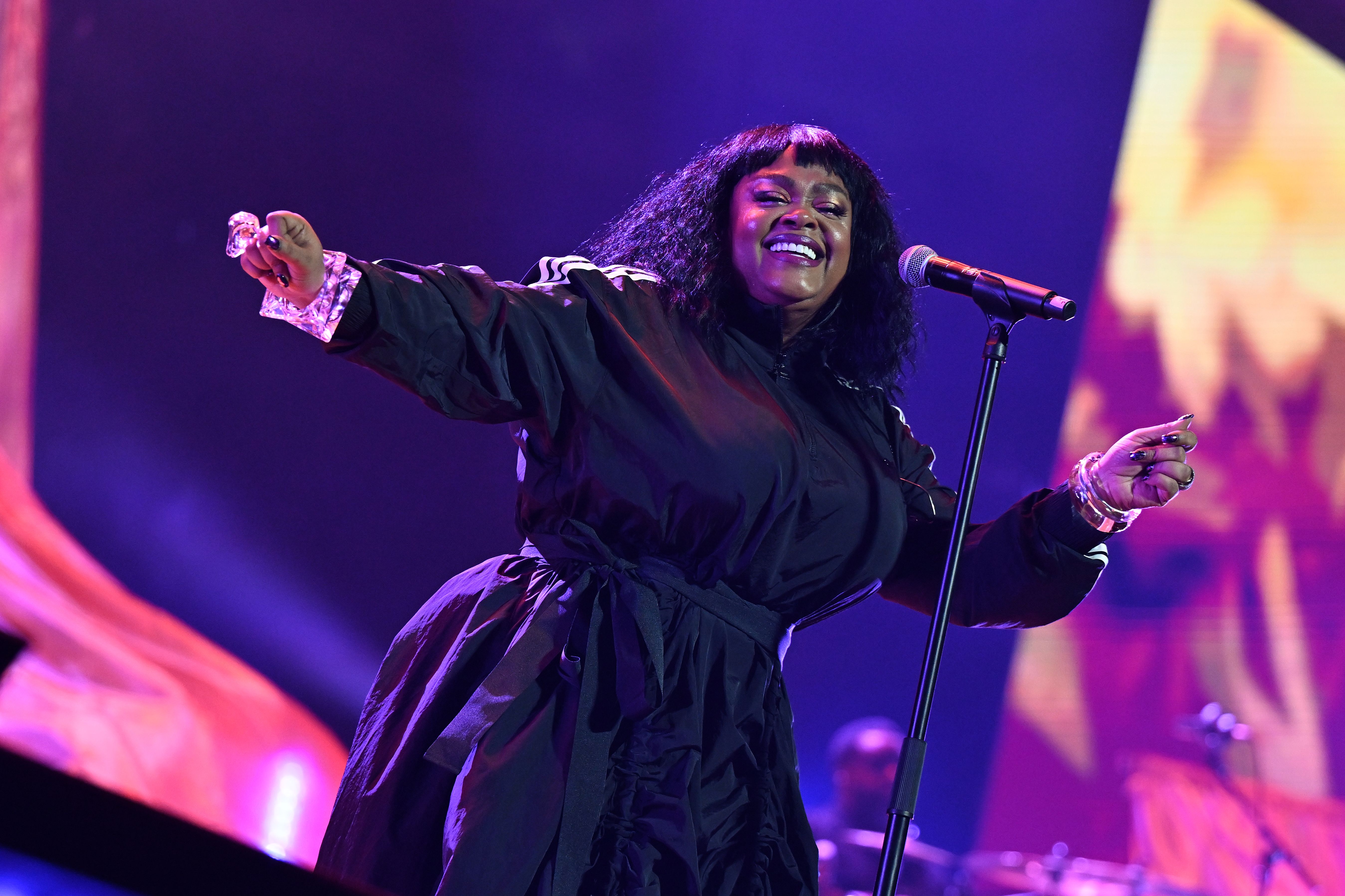 Singer in a black outfit with long sleeves and a belt performing on stage with a microphone, purple and orange lighting in the background, smiling and dancing.