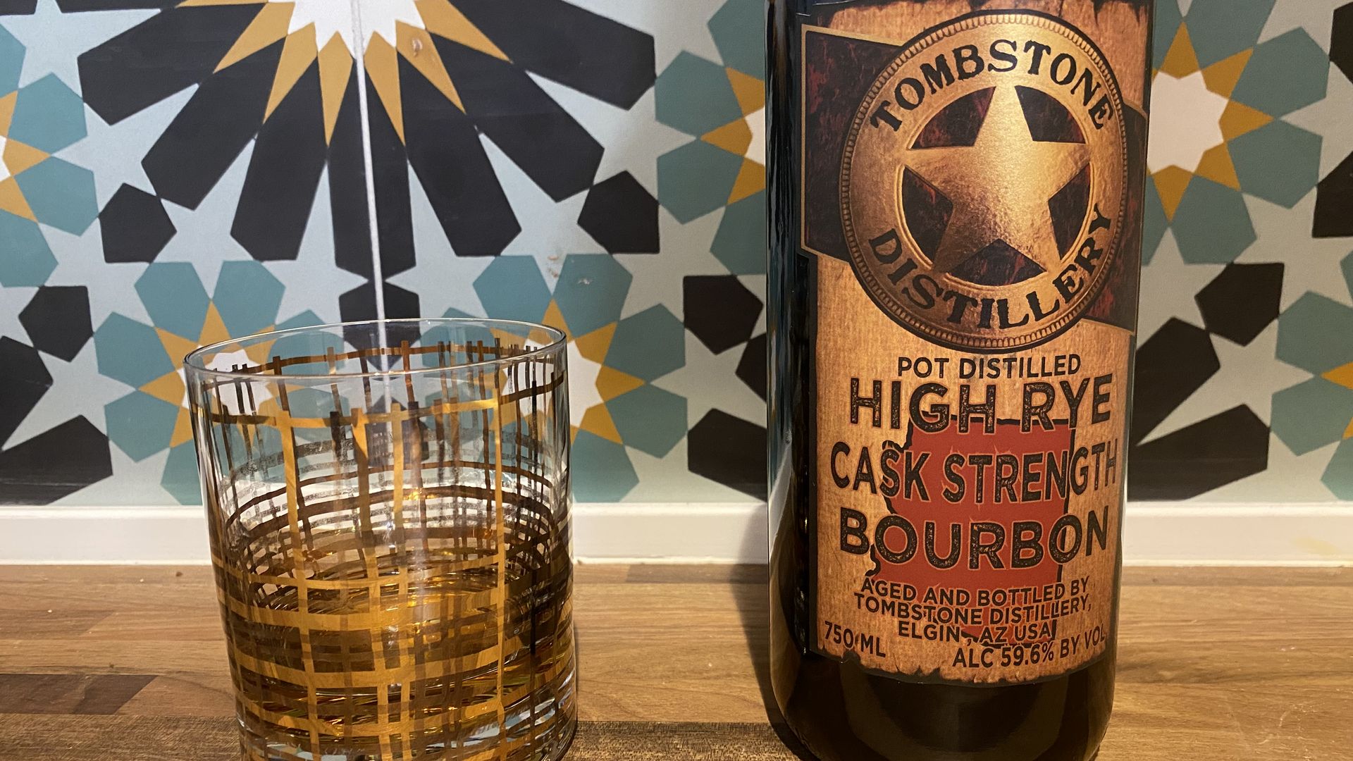 A clear whiskey glass with decorative gold lines next to a brown bourbon bottle in front of multicolored tile.  