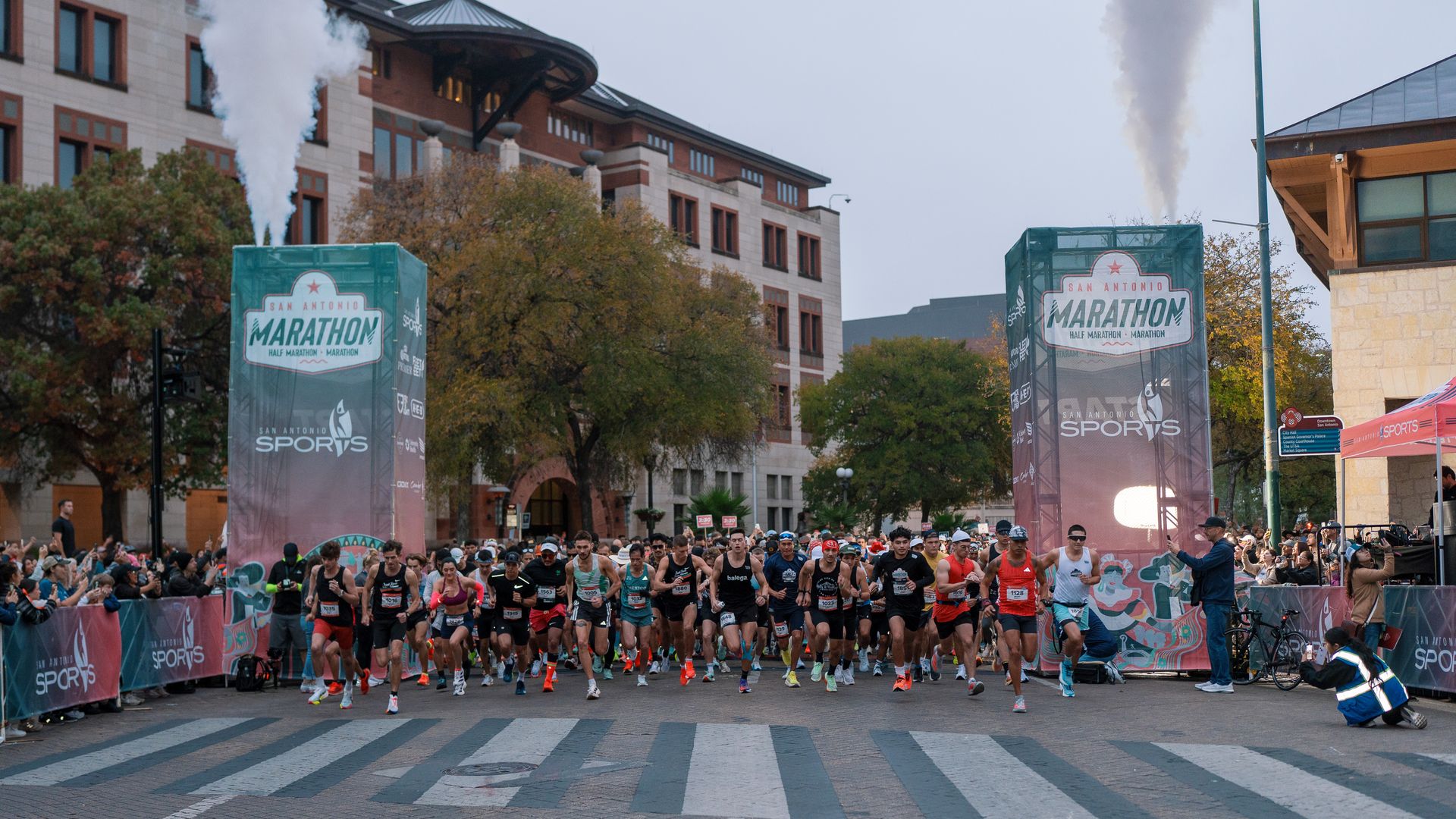 Runners at the San Antonio Marathon start, crowds along barriers, two green start banners, city buildings and trees in the background, steam rising from vents.