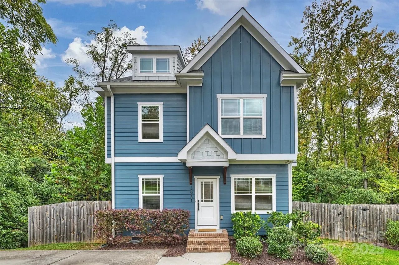 Two-story blue house with white trim, several windows, white front door, landscaped bushes, brick steps, wooden fence, and green trees under a partly cloudy sky.