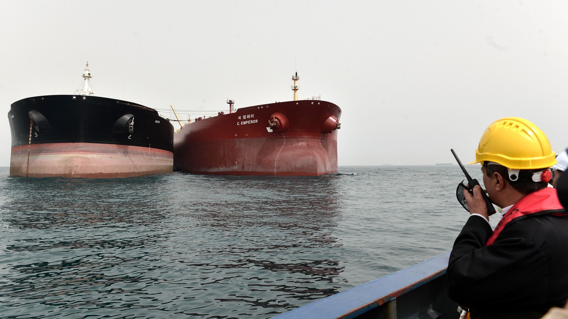 In this image, a man in a yellow hard hat speaks into a walkie talkie while looking out at two large tankers on the ocean.