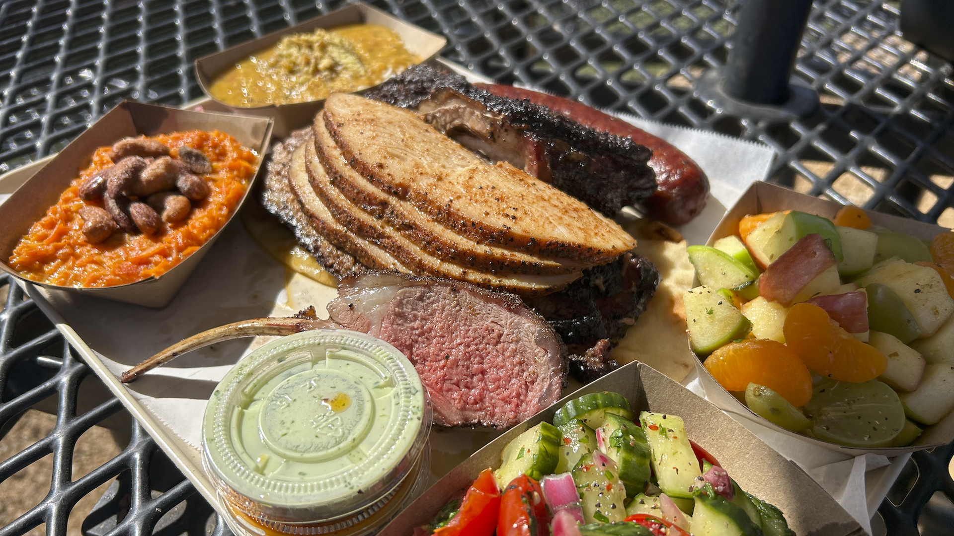 BBQ platter with sliced turkey, beef rib, sausage, sides of carrot salad with nuts, cucumber tomato salad, fruit salad, and two sauces on a metal outdoor table.