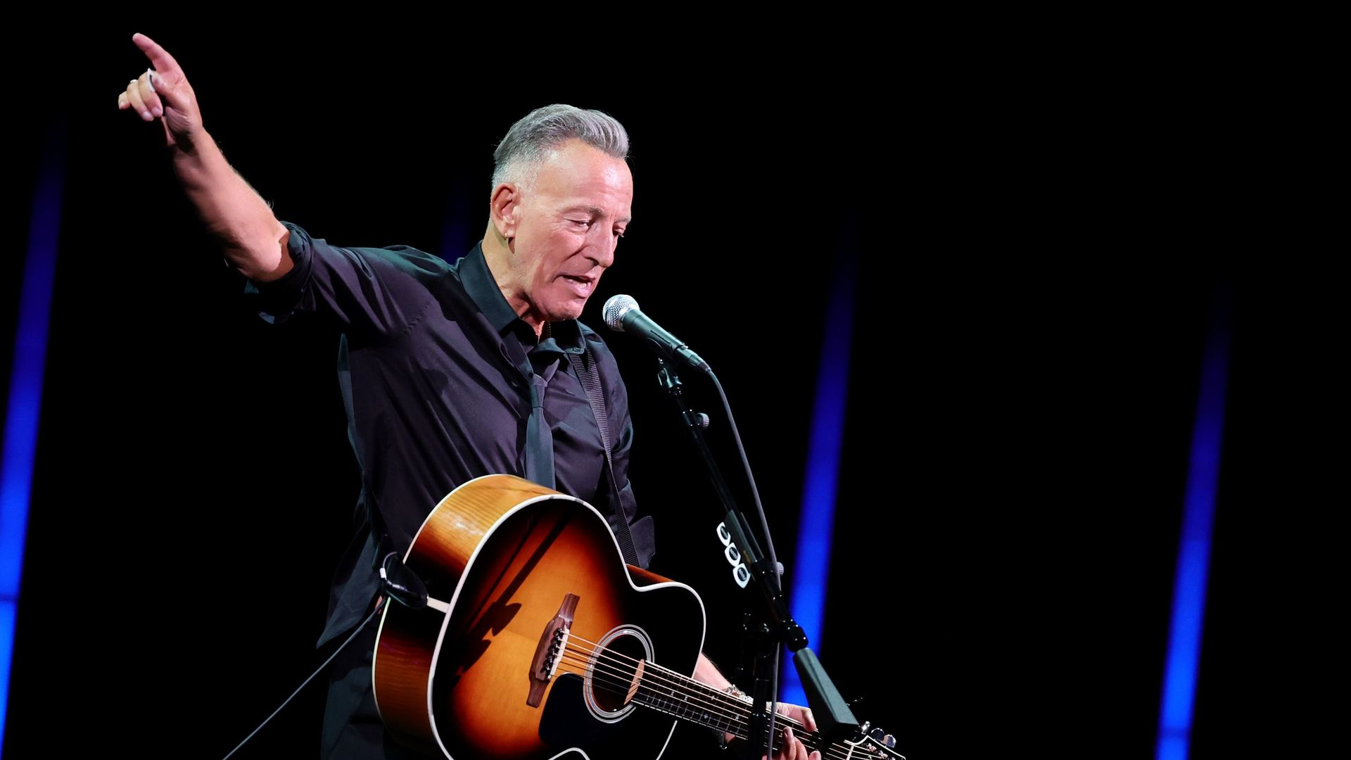 Bruce Springsteen wearing a black shirt and tie, holding a brown guitar and raising his hand in the air. 