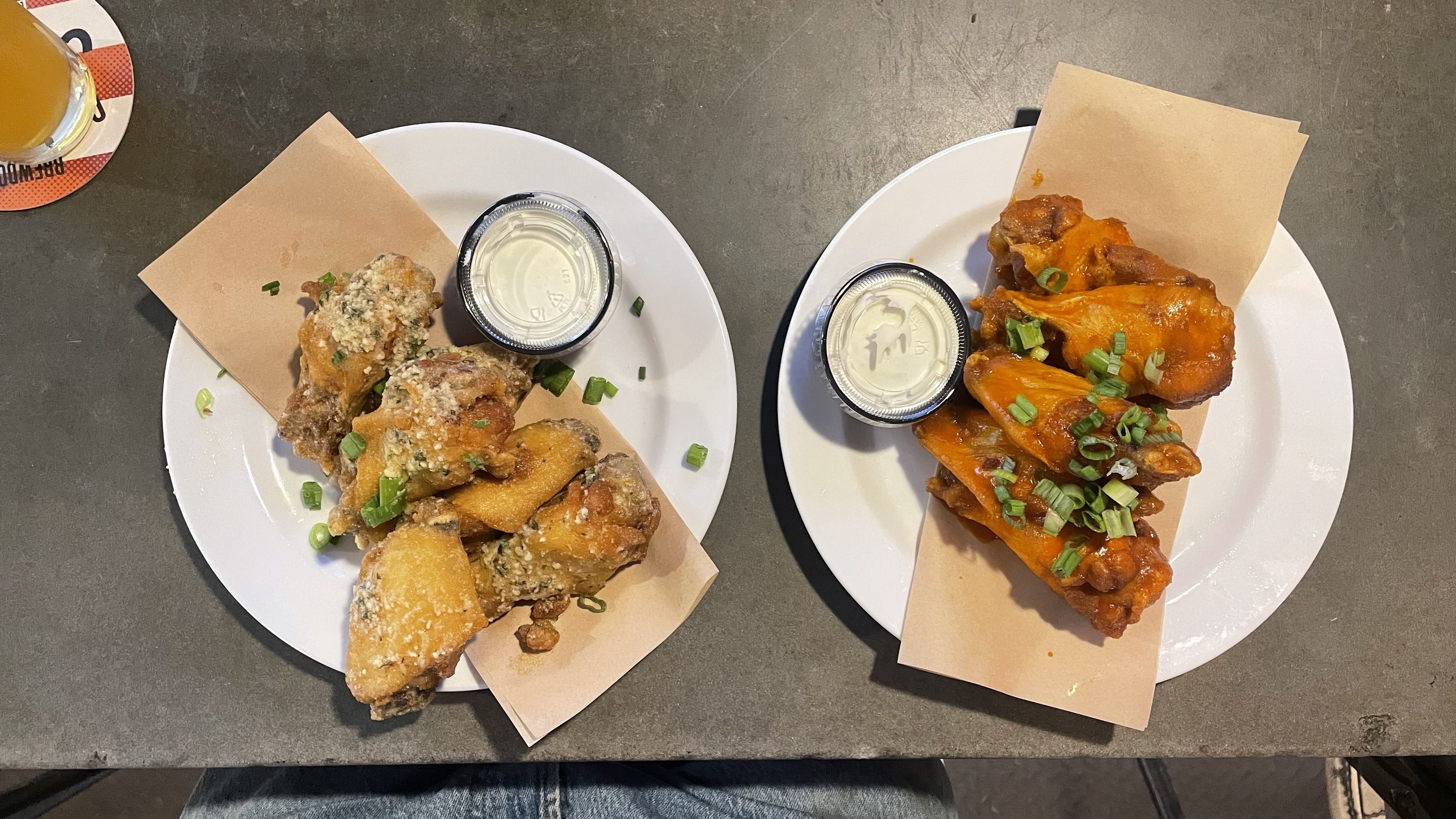 Two plates of chicken wings on a gray table. Left plate has parmesan garlic wings with dipping sauce; right plate has buffalo wings with green onion garnish and dipping sauce.