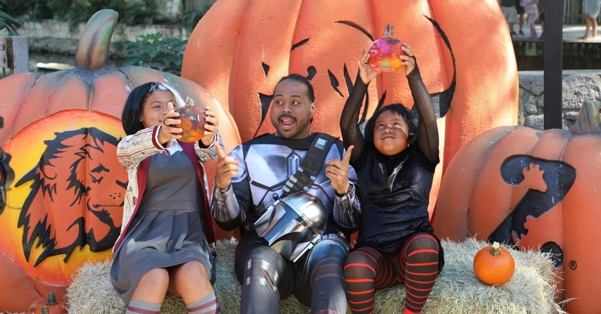 Two girls and a man sit on a stack of hay, looking at small pumpkins. 