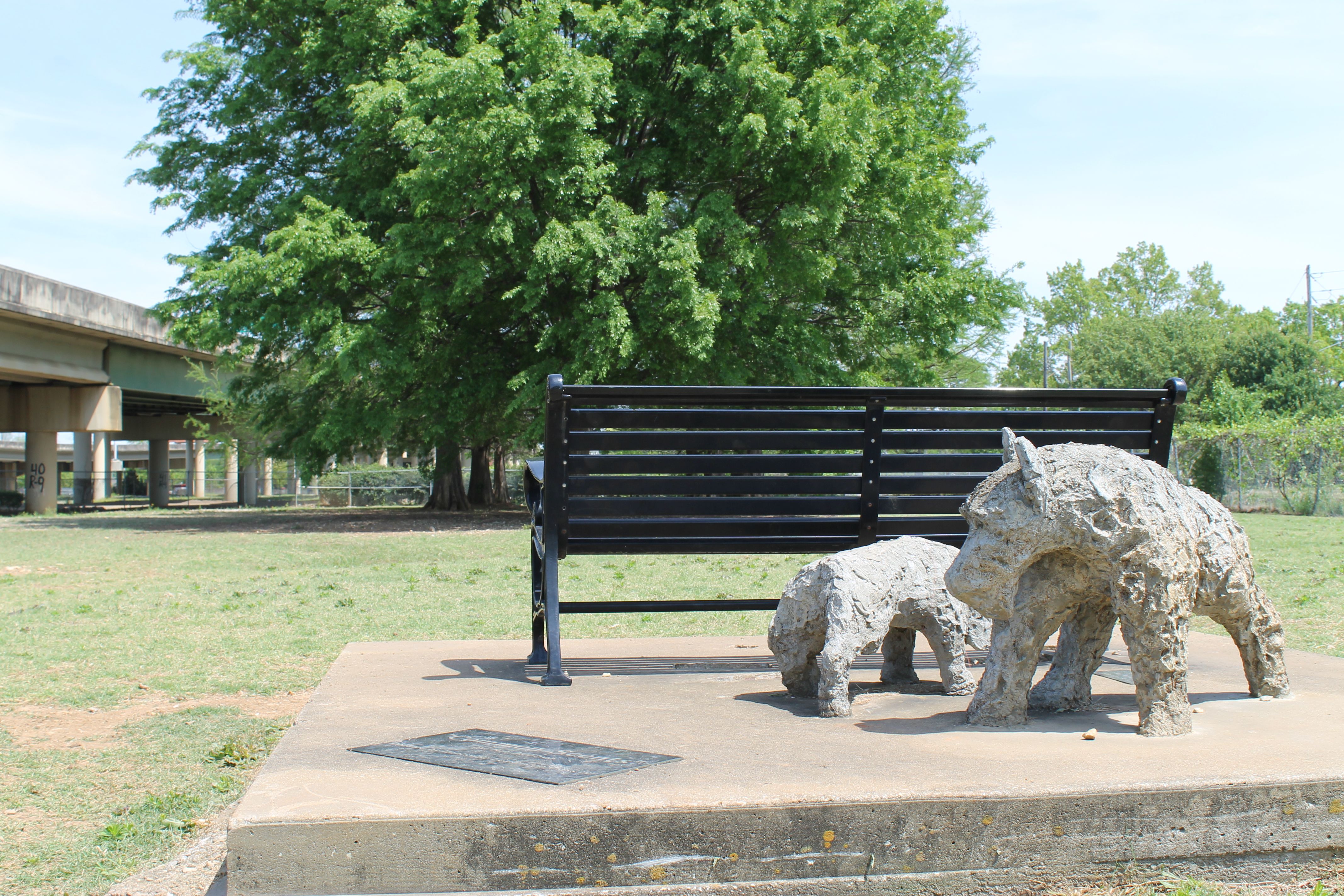 A black metal bench on a concrete platform in a sunny park, with two rough gray stone elephant sculptures nearby; a large leafy tree and an overpass are visible in the background.