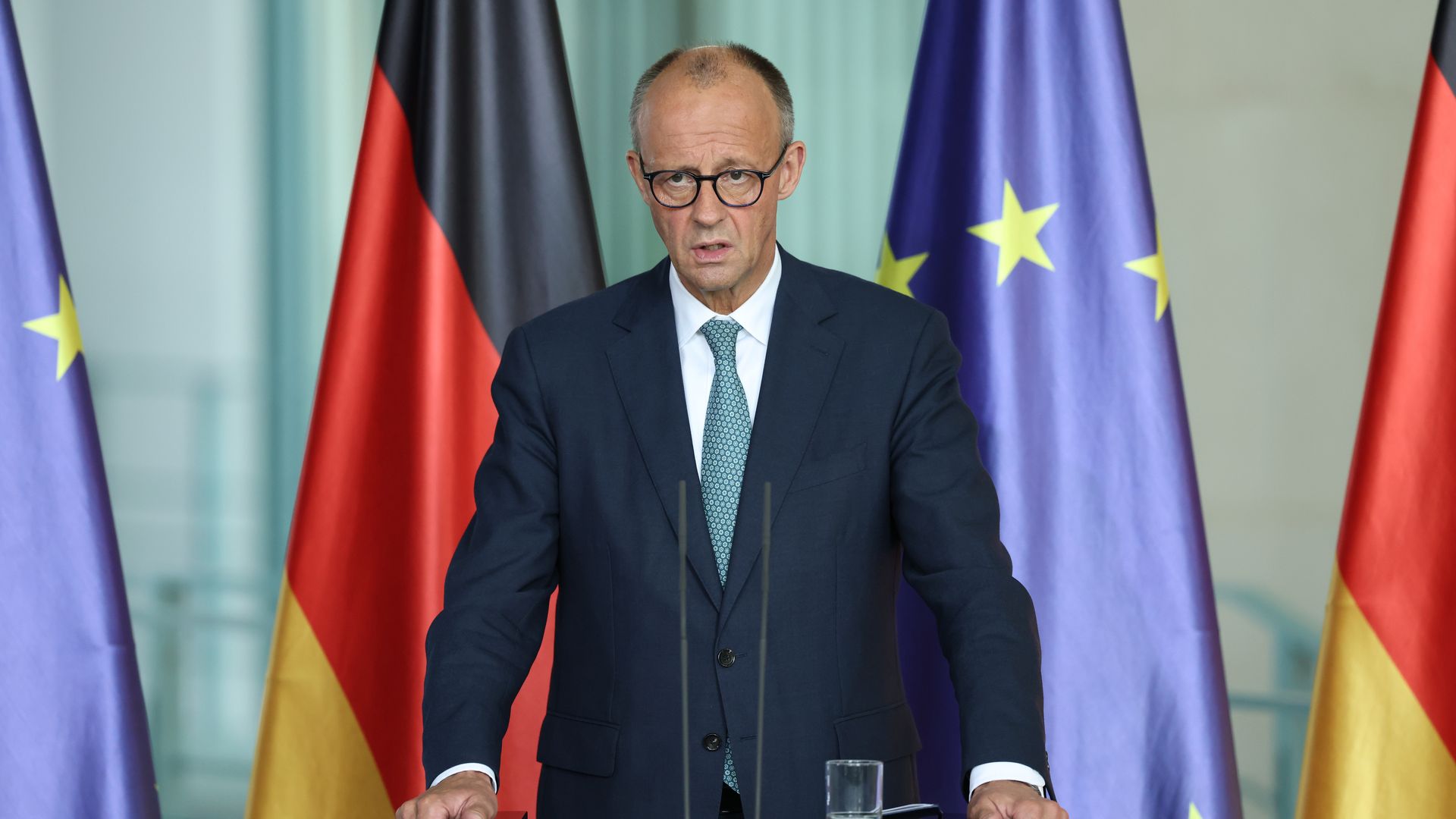 Man in dark suit and glasses standing at a podium with German and European Union flags behind him, speaking in a formal setting.