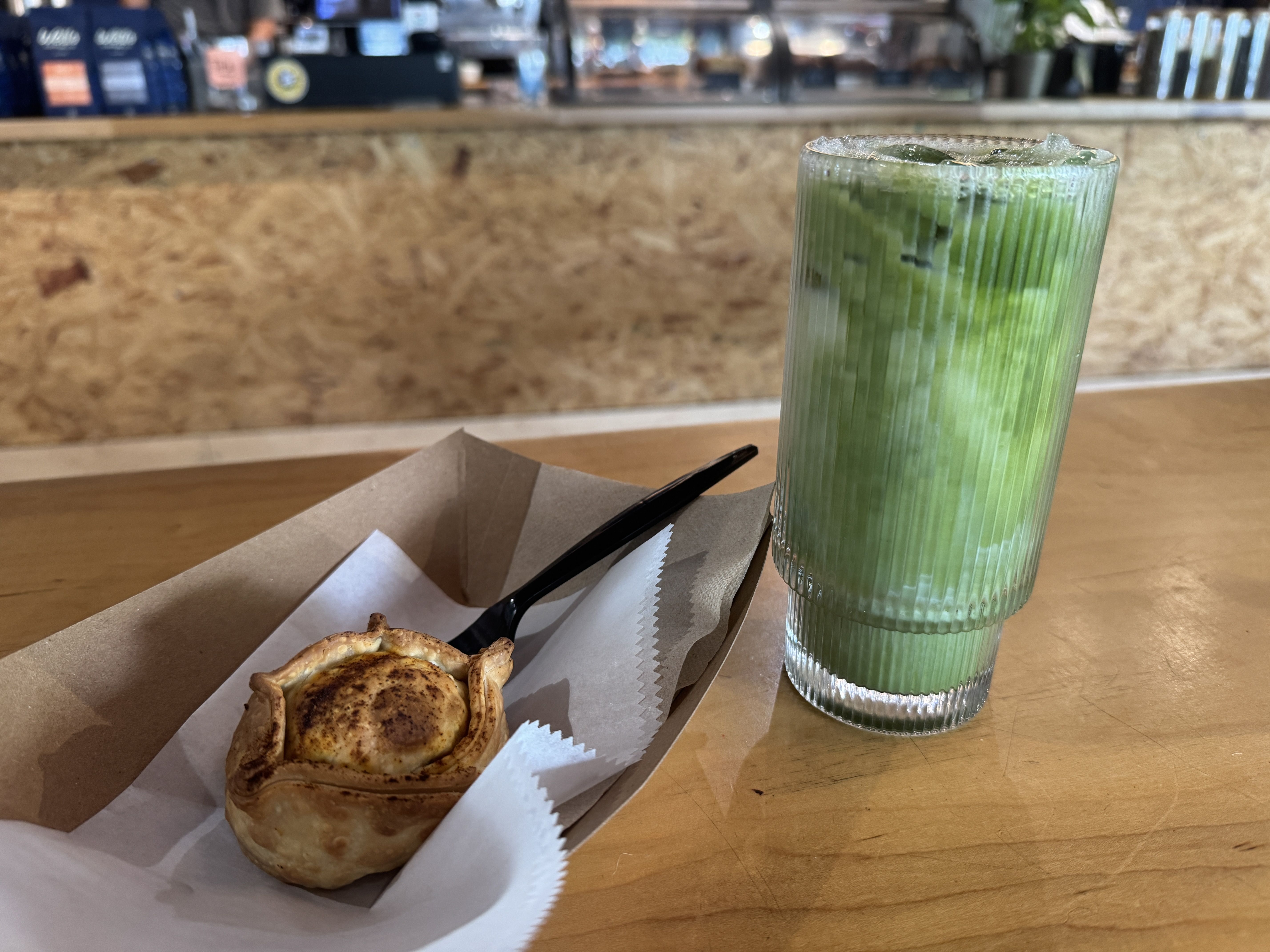 A tall ribbed glass with a bright green iced drink sits on a wooden table beside a cinnamon roll in a parchment liner; a blurred cafe counter is visible in the background.