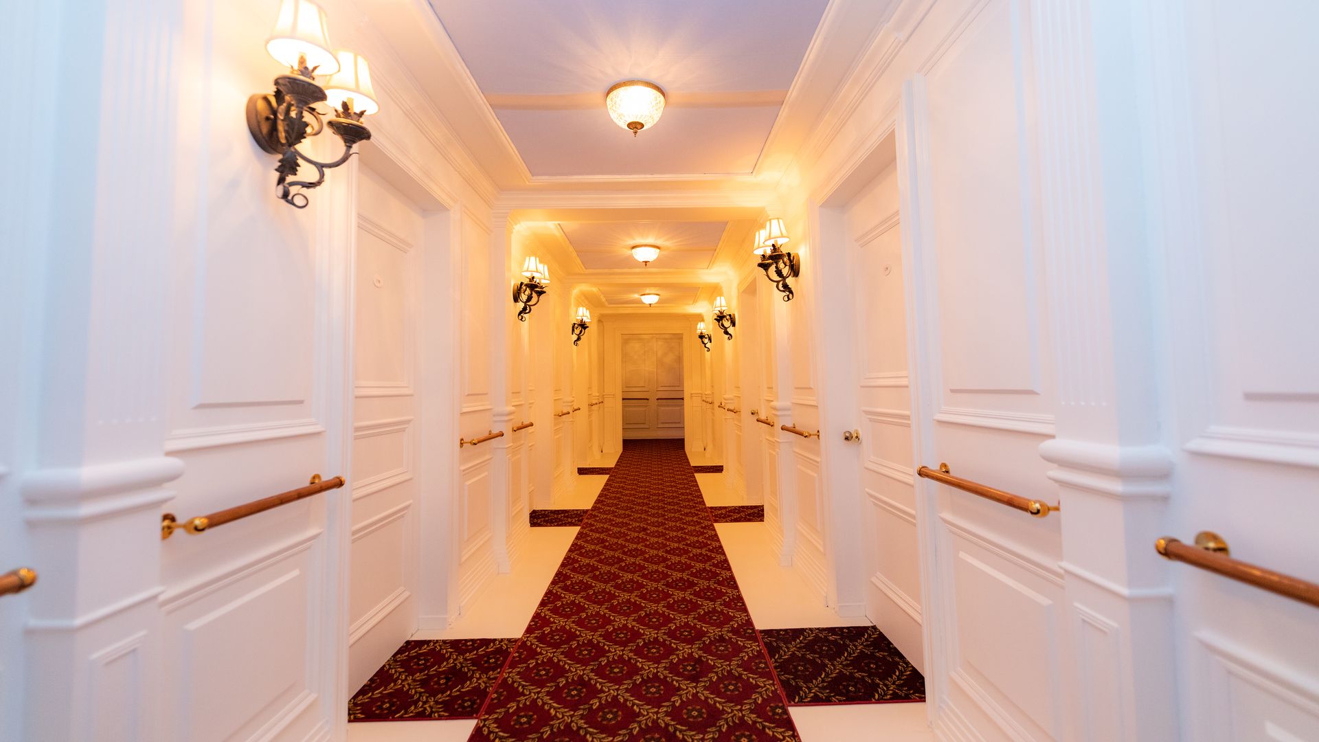 Long, elegant hotel hallway with white walls, ornate wall sconces, ceiling lights, and a red patterned carpet runner extending to double doors at the end.