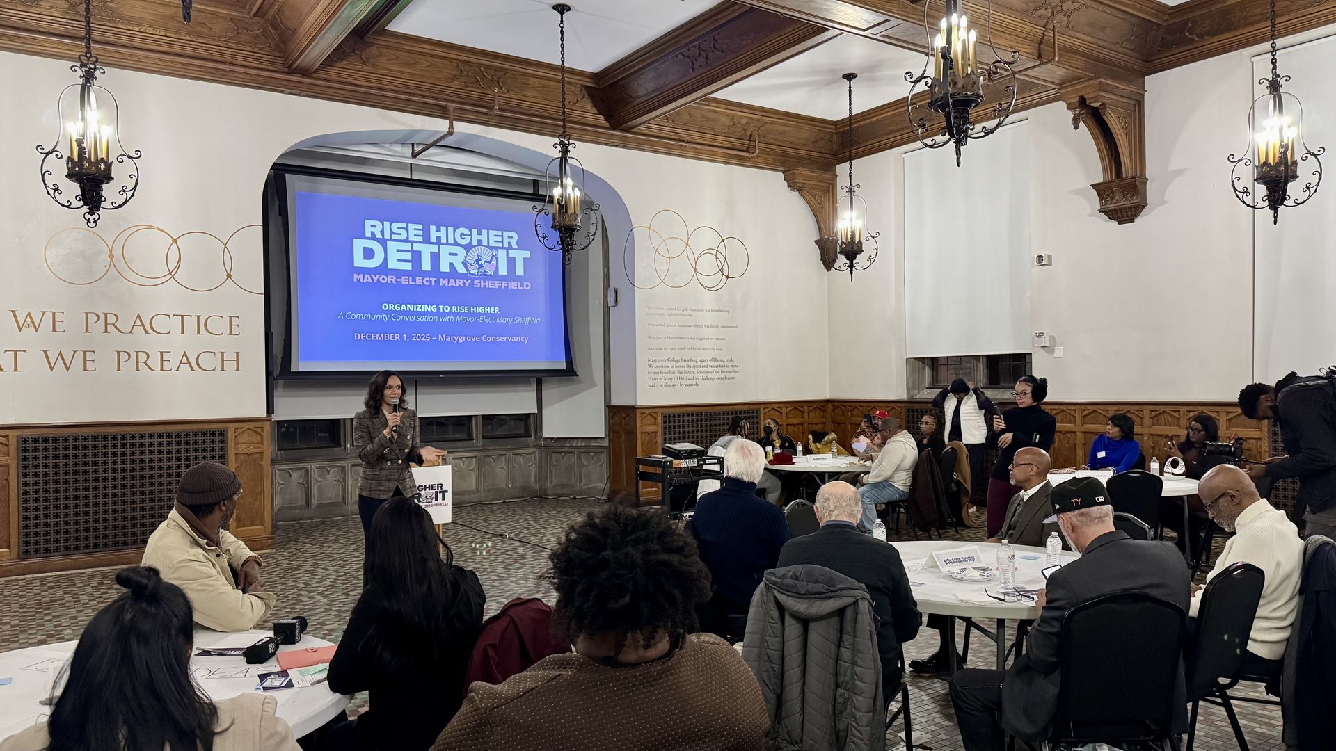 Sheffield speaks to an audience in a wood-paneled room with chandeliers. A blue screen behind her reads "Rise Higher Detroit Mayor-Elect Mary Sheffield"