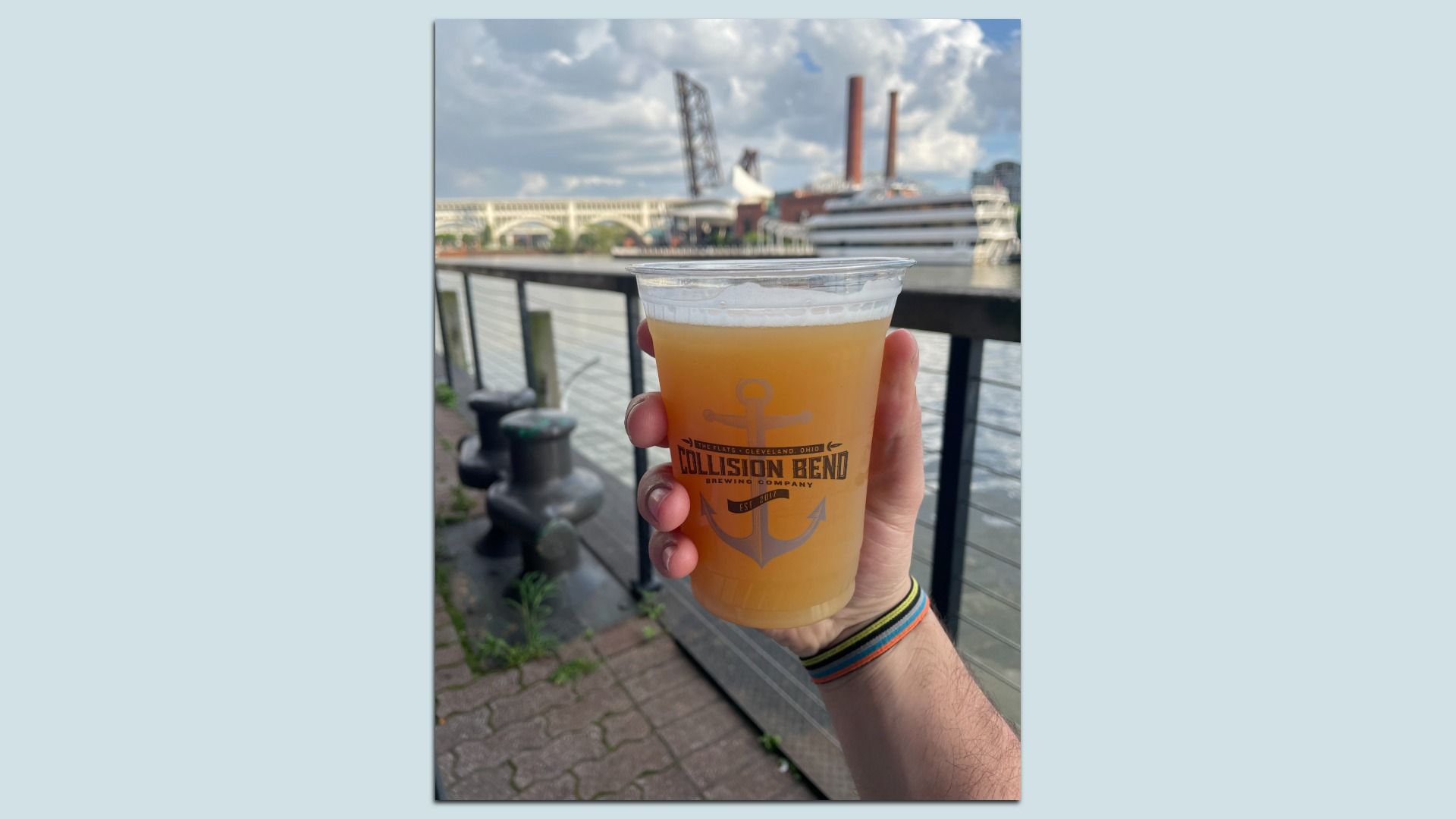 Hand holding beer on riverfront patio, bridge in background