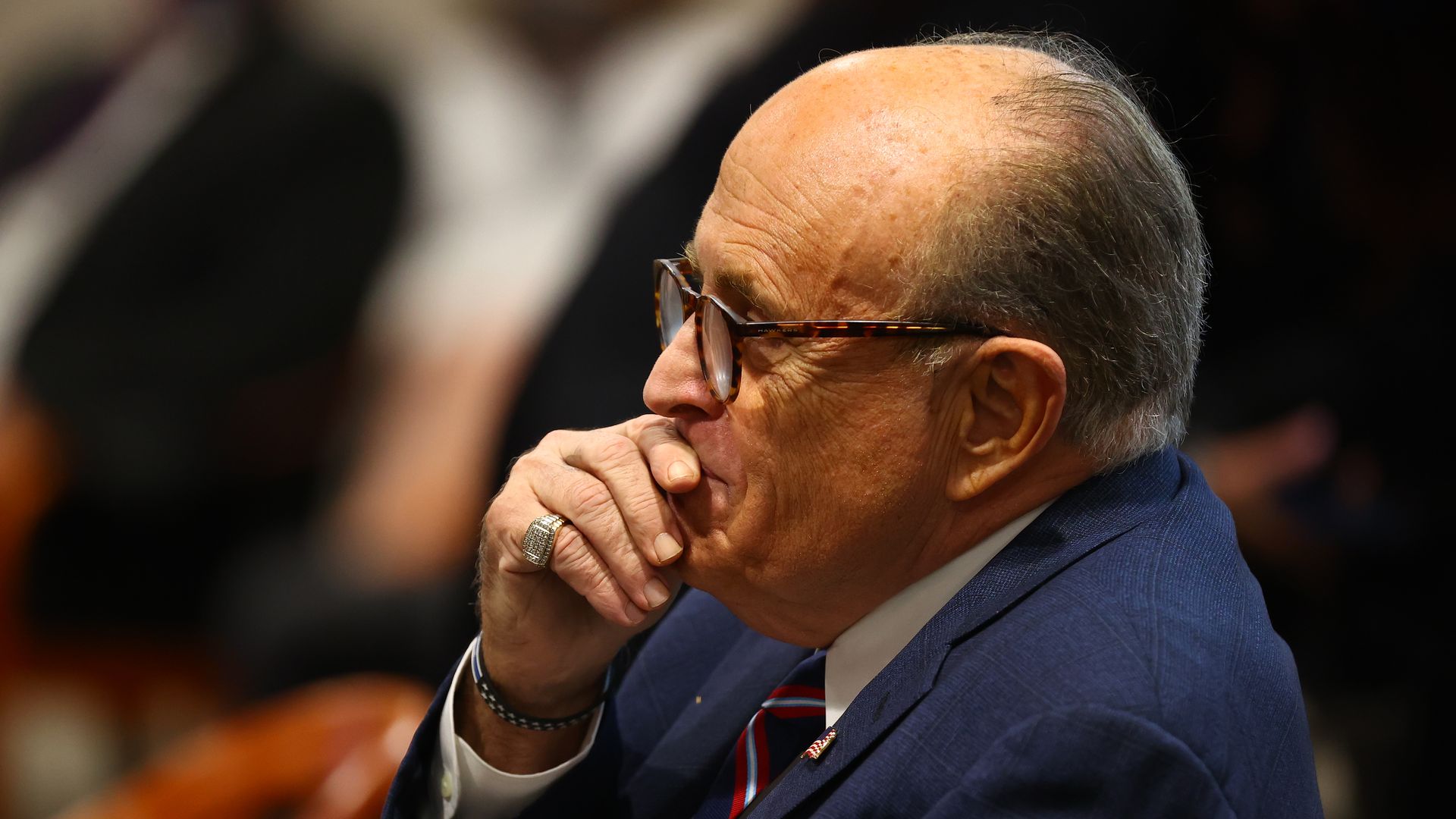 Rudy Giuliani wears a blue suit and is photographed from his side profile. His hand is over his mouth. 