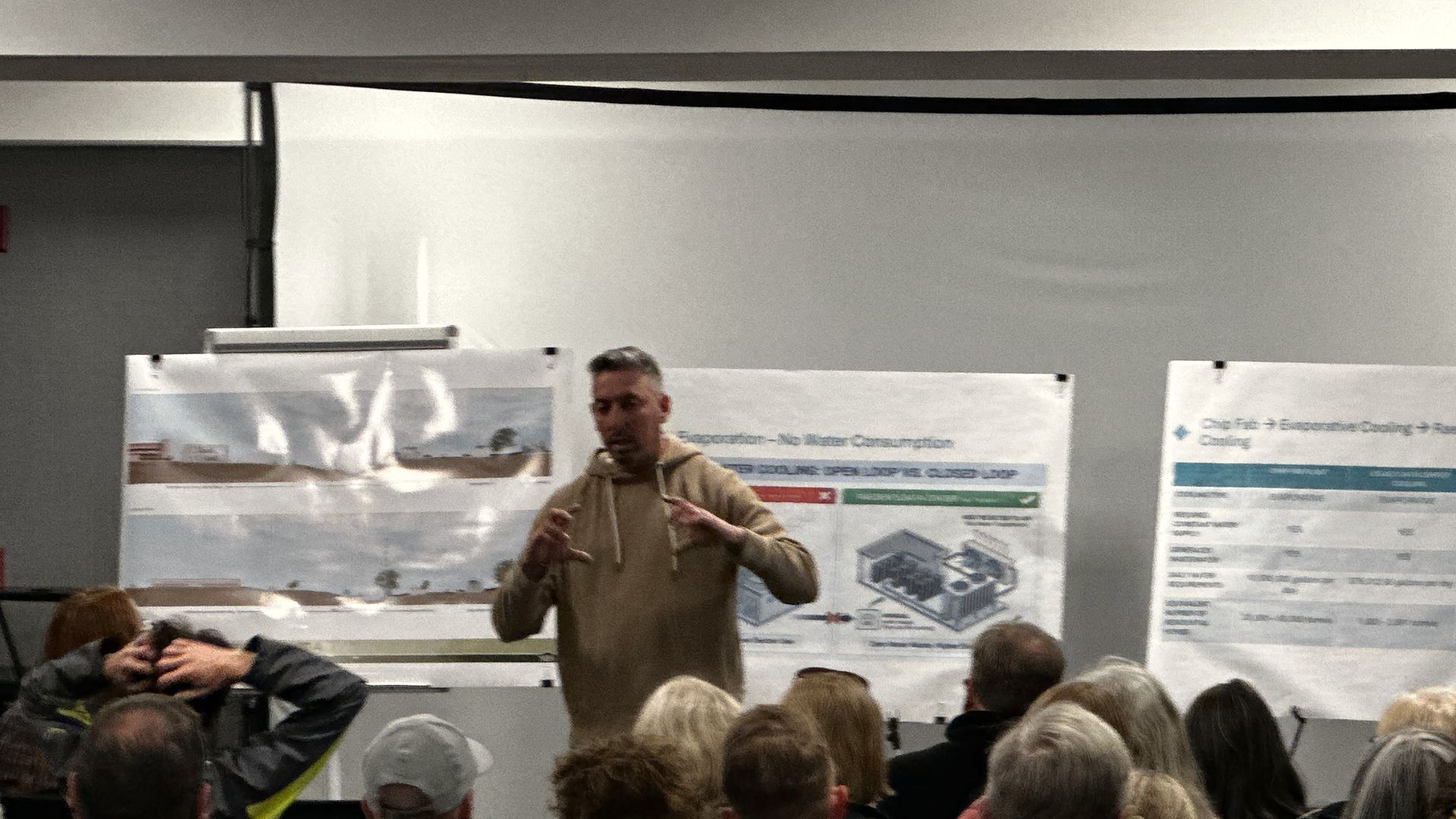 A man in a beige hoodie stands at the front of a conference room, gesturing as he speaks to a seated audience. Large posters with diagrams hang behind him.