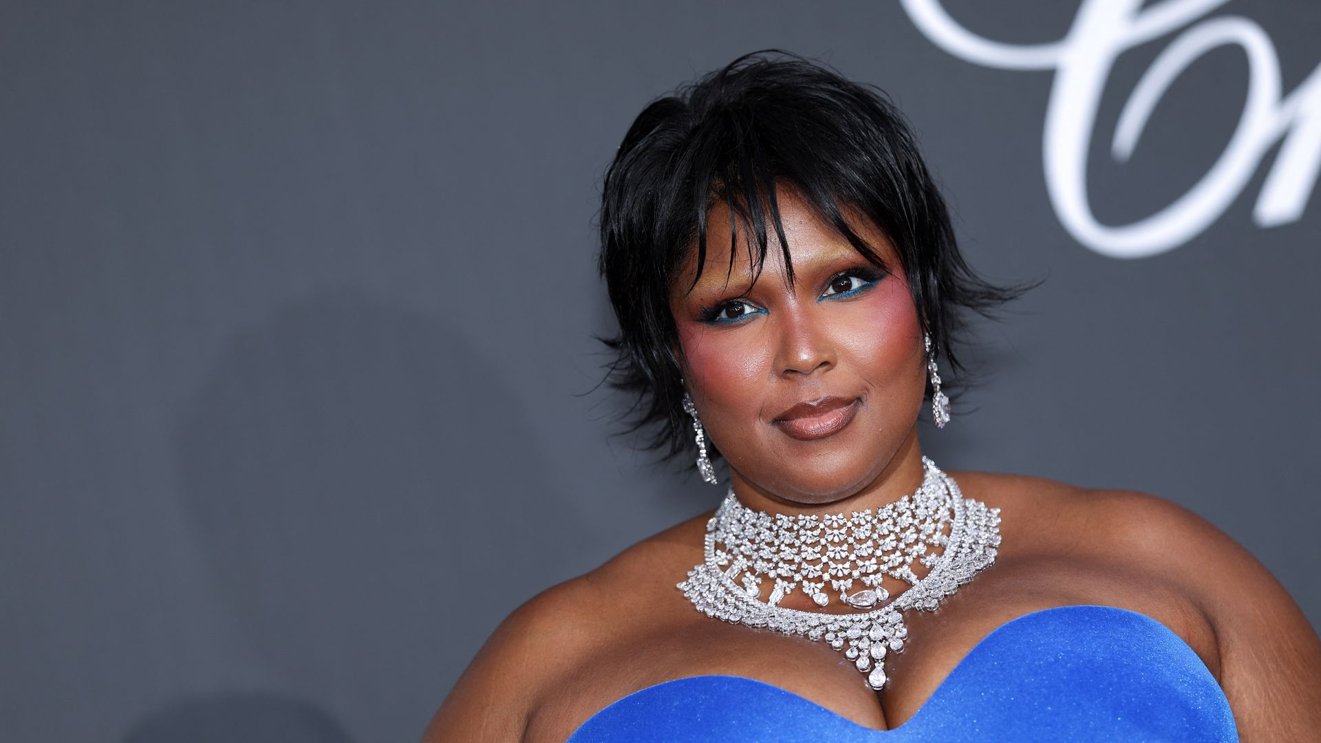 Close-up of a woman with short black hair, bold makeup (blue eyeshadow and pink blush), wearing a diamond necklace and earrings, in a blue off-the-shoulder gown against a gray backdrop.