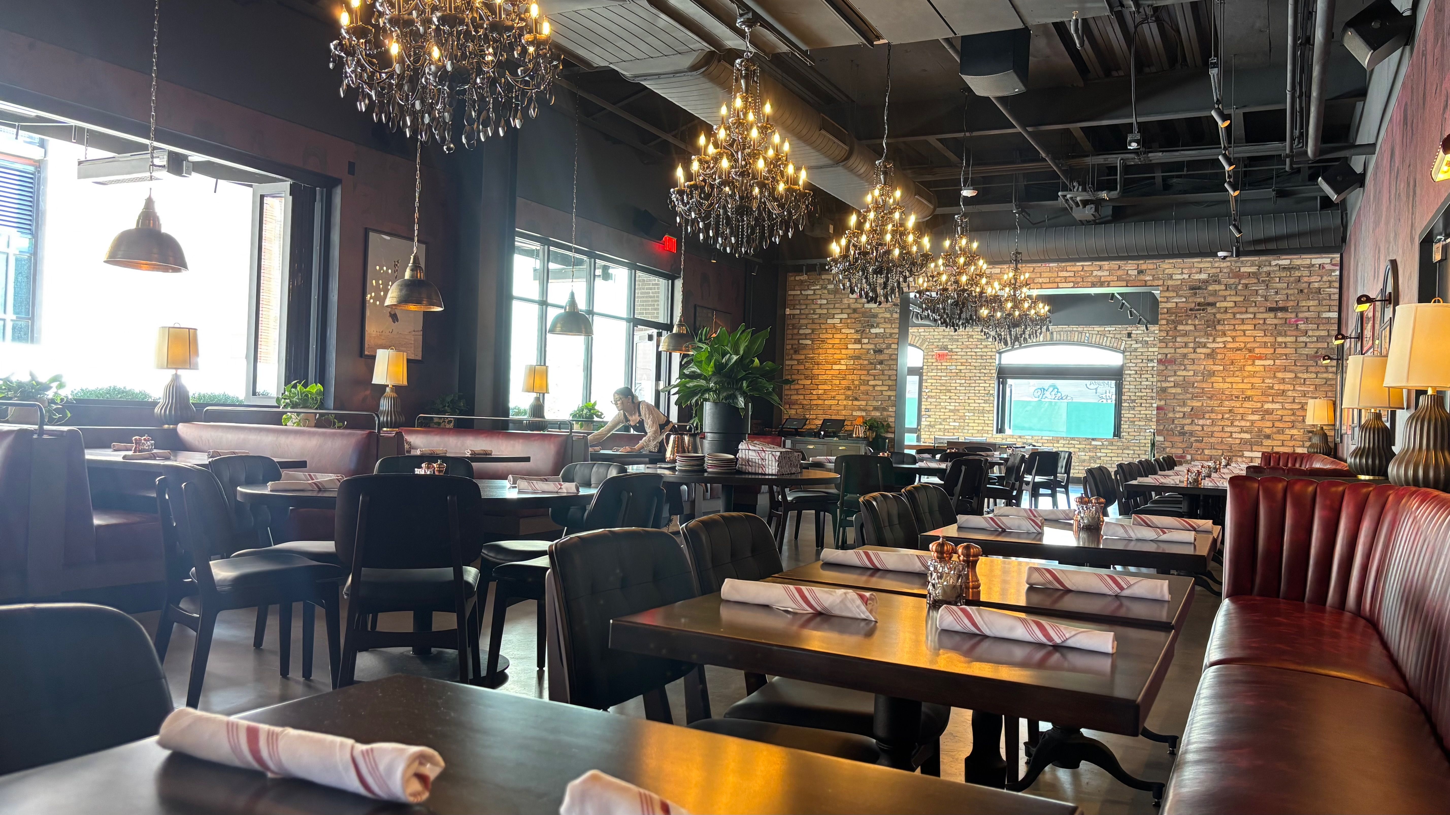 A stylish restaurant interior with exposed brick walls, crystal chandeliers, and rows of tables set with napkins. Bright daylight from the left windows and a server tending a table near the center.