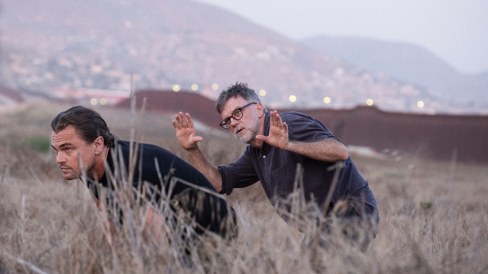 Director-writer-producer Paul Thomas Anderson and Leonardo DiCaprio crouch in dry grass; one wears black and leans forward, the other in glasses and dark shirt gestures with raised hands. Hills and blurred lights in the background.