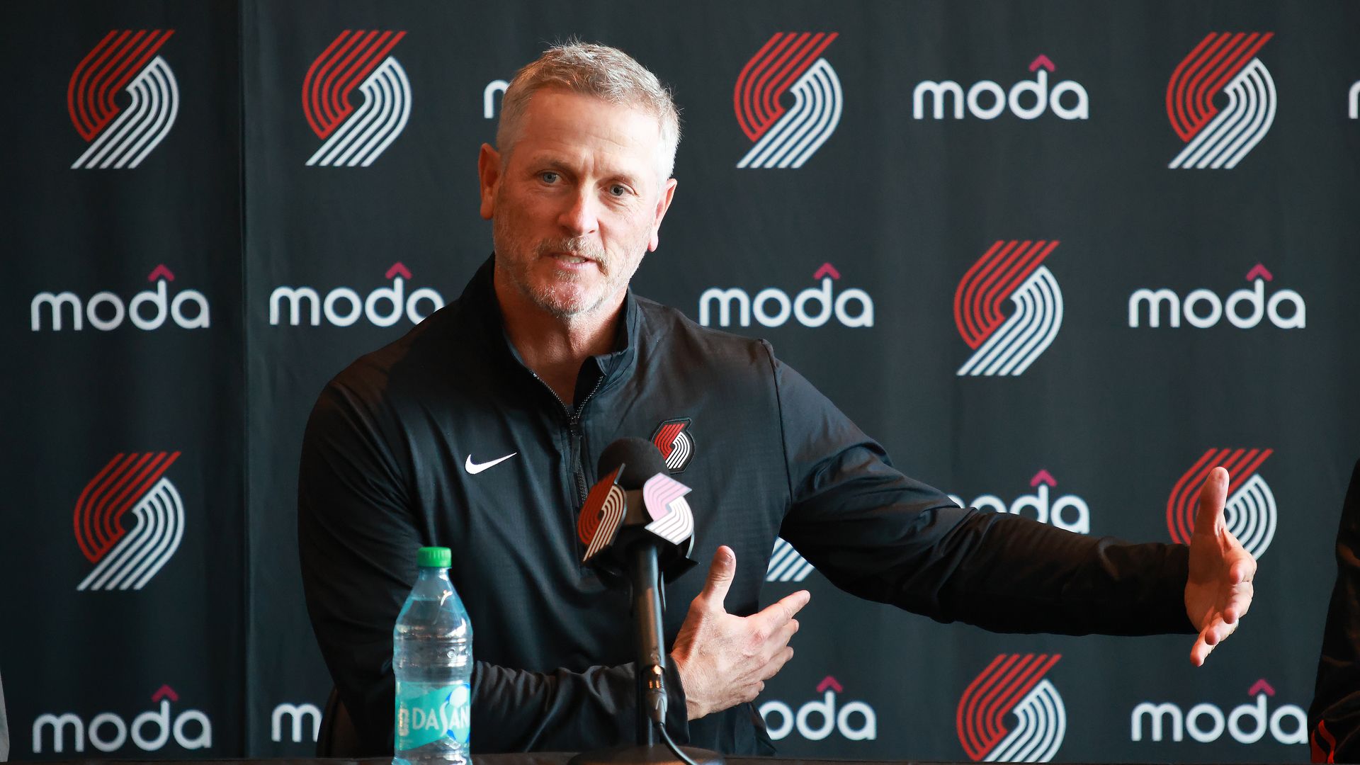 Blazers owner Tom Dundon speaks at a press conference, in front of a black backdrop with red and white modo logos; he gestures with his hands, while a microphone and water bottle sit on the table.