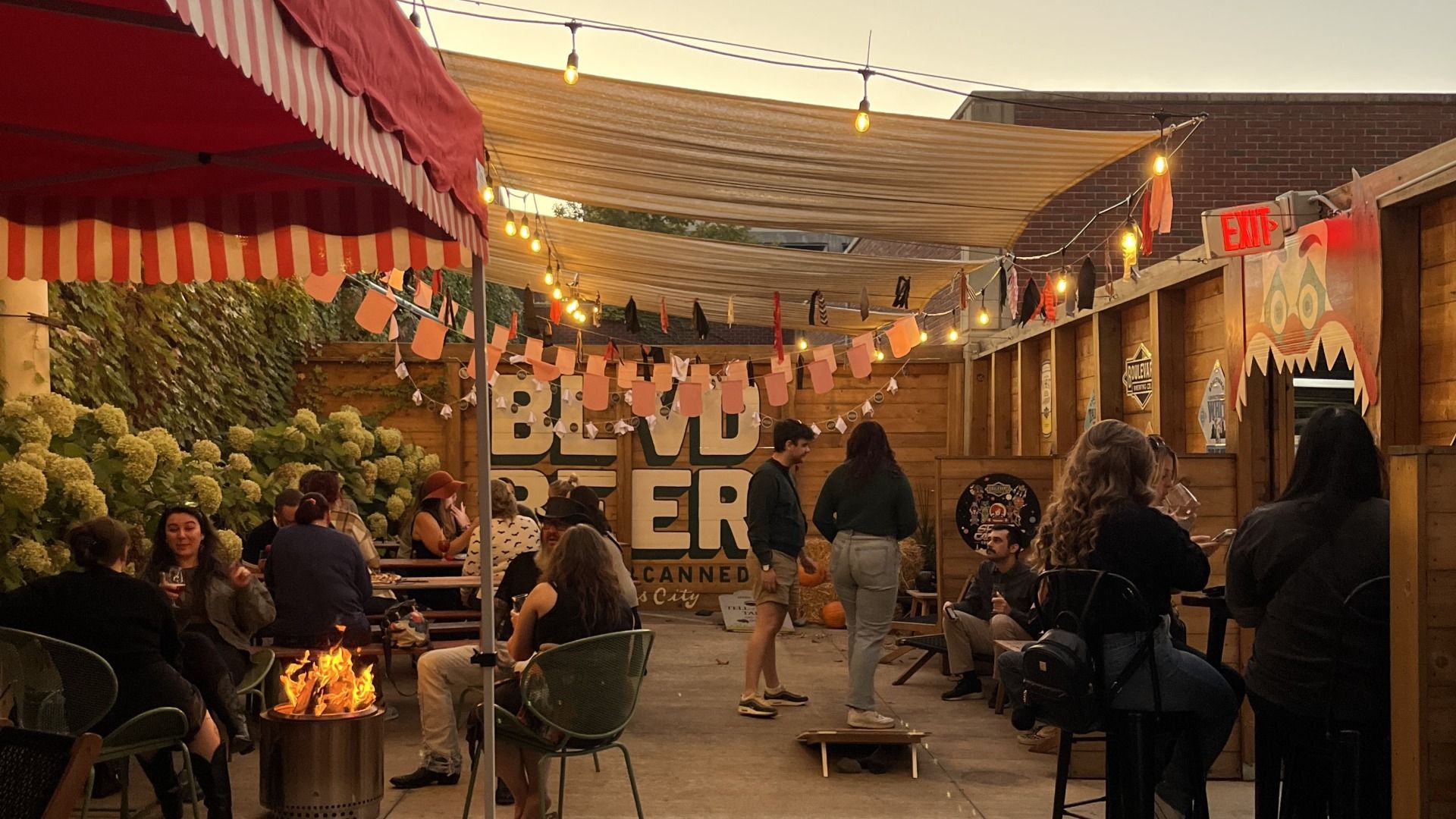 Outdoor evening gathering with people sitting, standing, and chatting under yellow string lights and striped awnings, near a fire pit and wooden walls with signs and decorations.