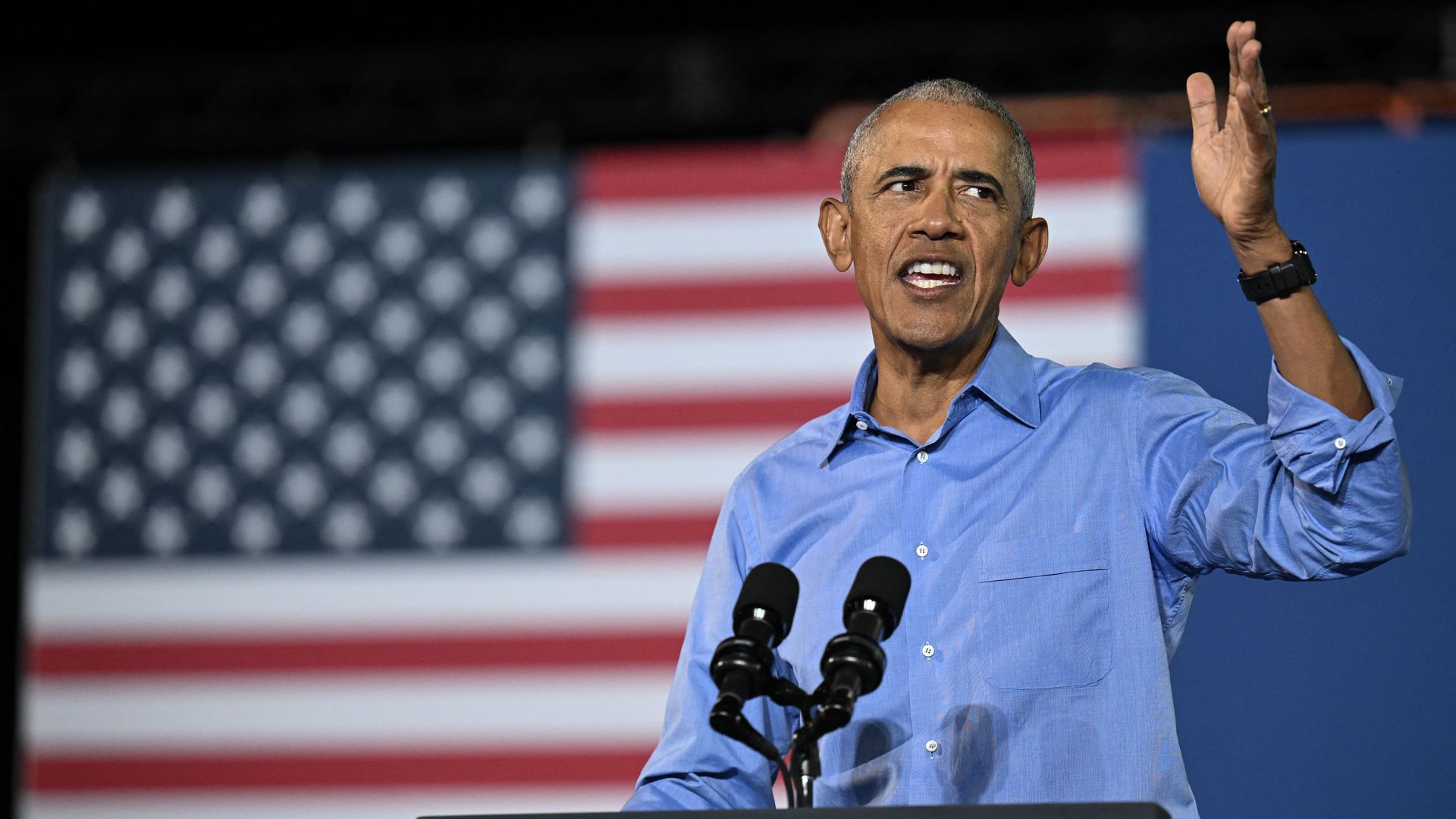 A man wearing a blue collared, long-sleeved buttoned shirt speaks into two microphones attached to a podium. His left hand is in the air and an American flag is in the background