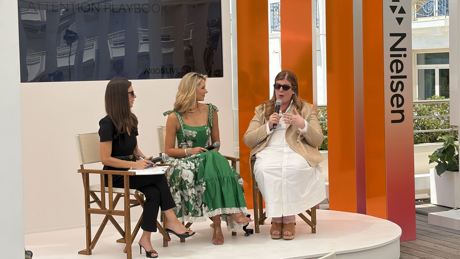 Panel discussion at a Nielsen event: a large Nielsen sign, dark screen with the event title, Axios' Sara Fischer, ESPN host Laura Rutledge and Rita Ferro, president of global advertising at Disney seated on stage; orange pillars line the outdoor backdrop.