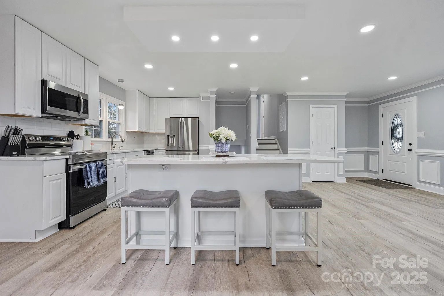 Bright, modern kitchen with white cabinets, stainless steel appliances, large island with three gray cushioned stools, light wood floors, and soft gray walls with white trim.