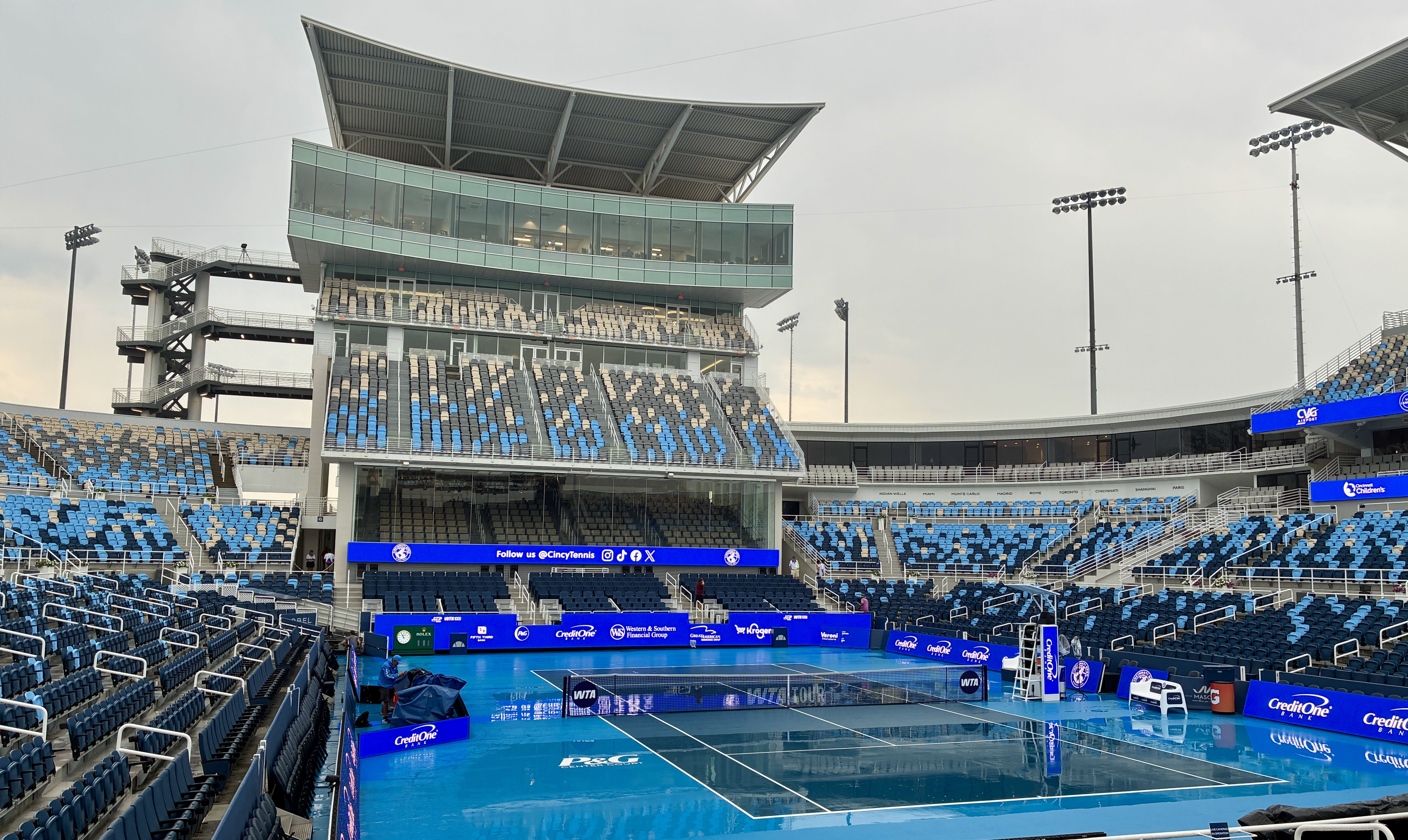 Wide view of a modern tennis stadium with a blue court, glass-fronted control tower, tiered blue and gray seating, and tall floodlights; banners display "Cincy Tennis".