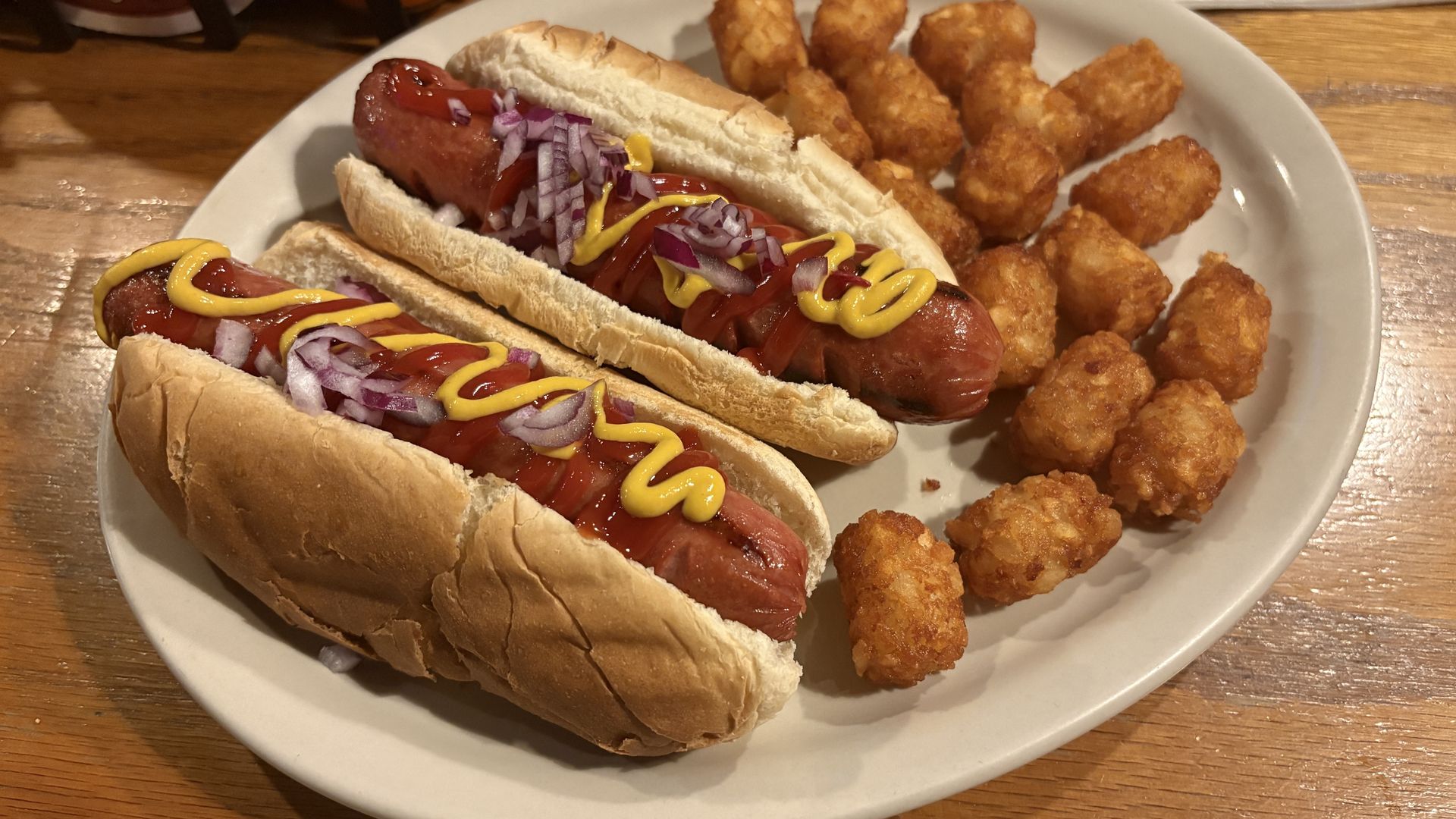Plate with two hot dogs in buns topped with ketchup, mustard, and chopped red onions, plus a cluster of golden fried nugget bites on a wooden table.