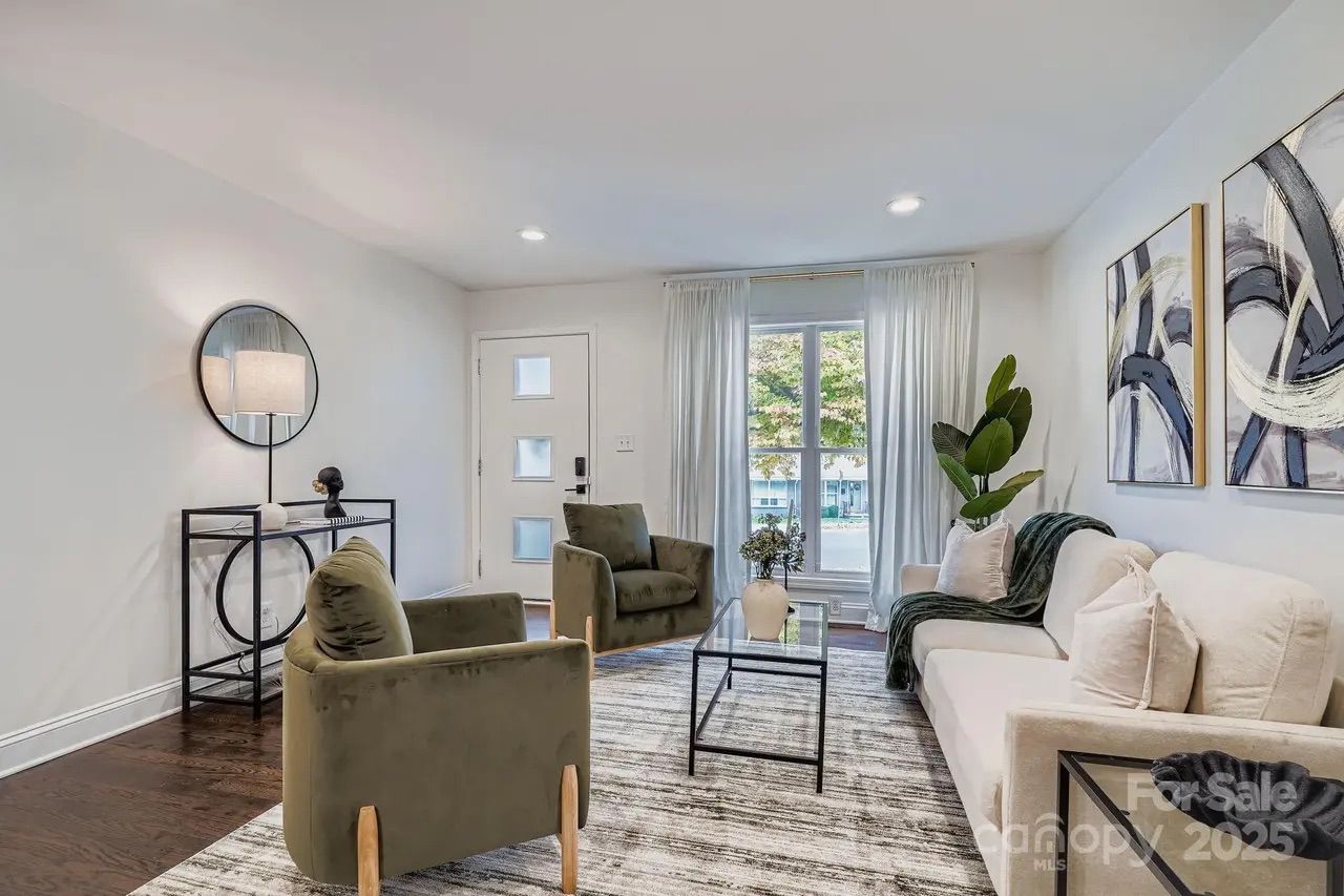 Bright living room with white walls, olive green chairs, beige sofa, glass coffee table, black metal console table with round mirror, abstract art on wall, large window with white curtains.