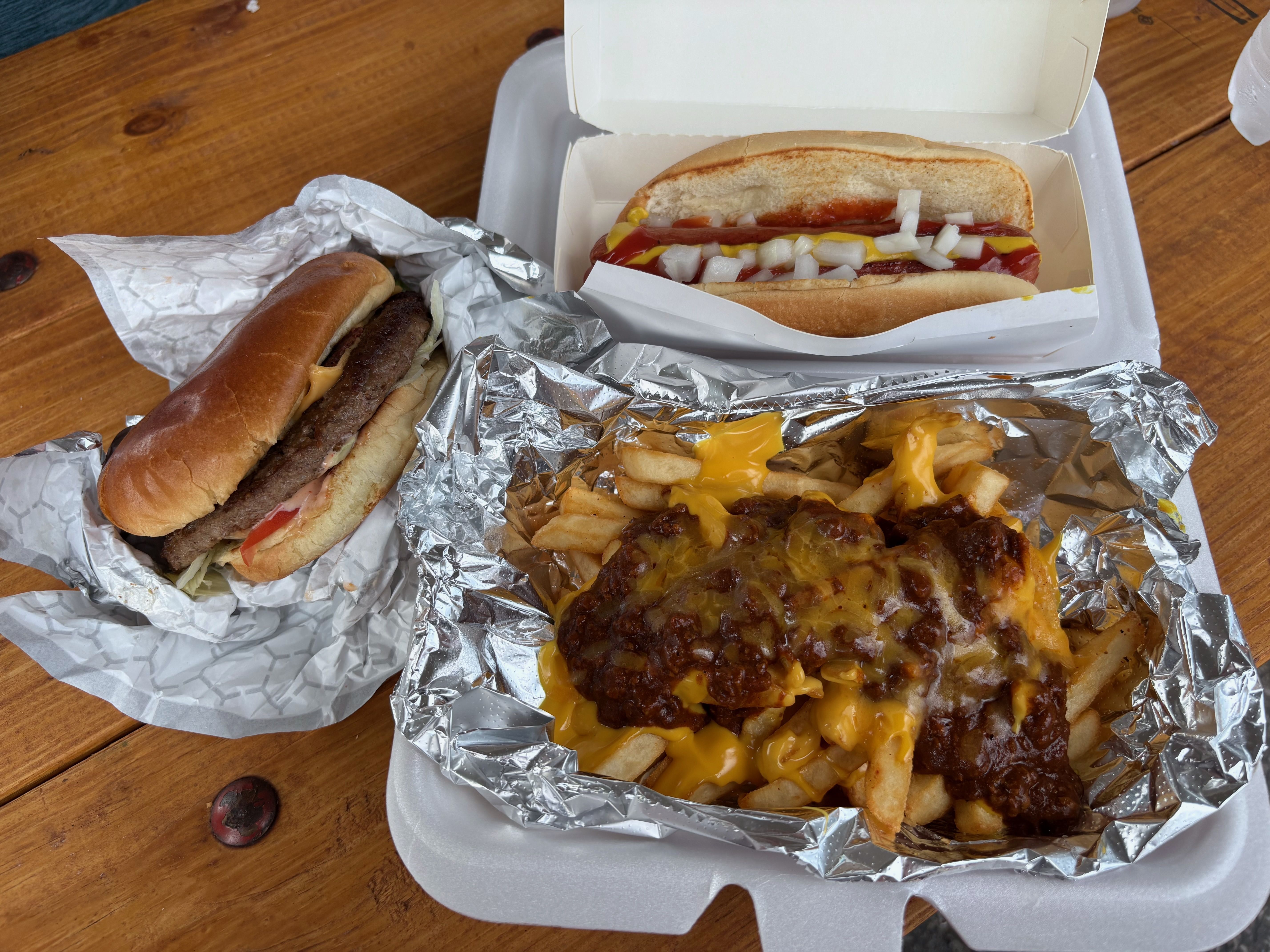 Top-down view of fast-food on a wooden table: hot dogs with onions in a white takeout box and foil-wrapped fries covered in chili and melted cheese.