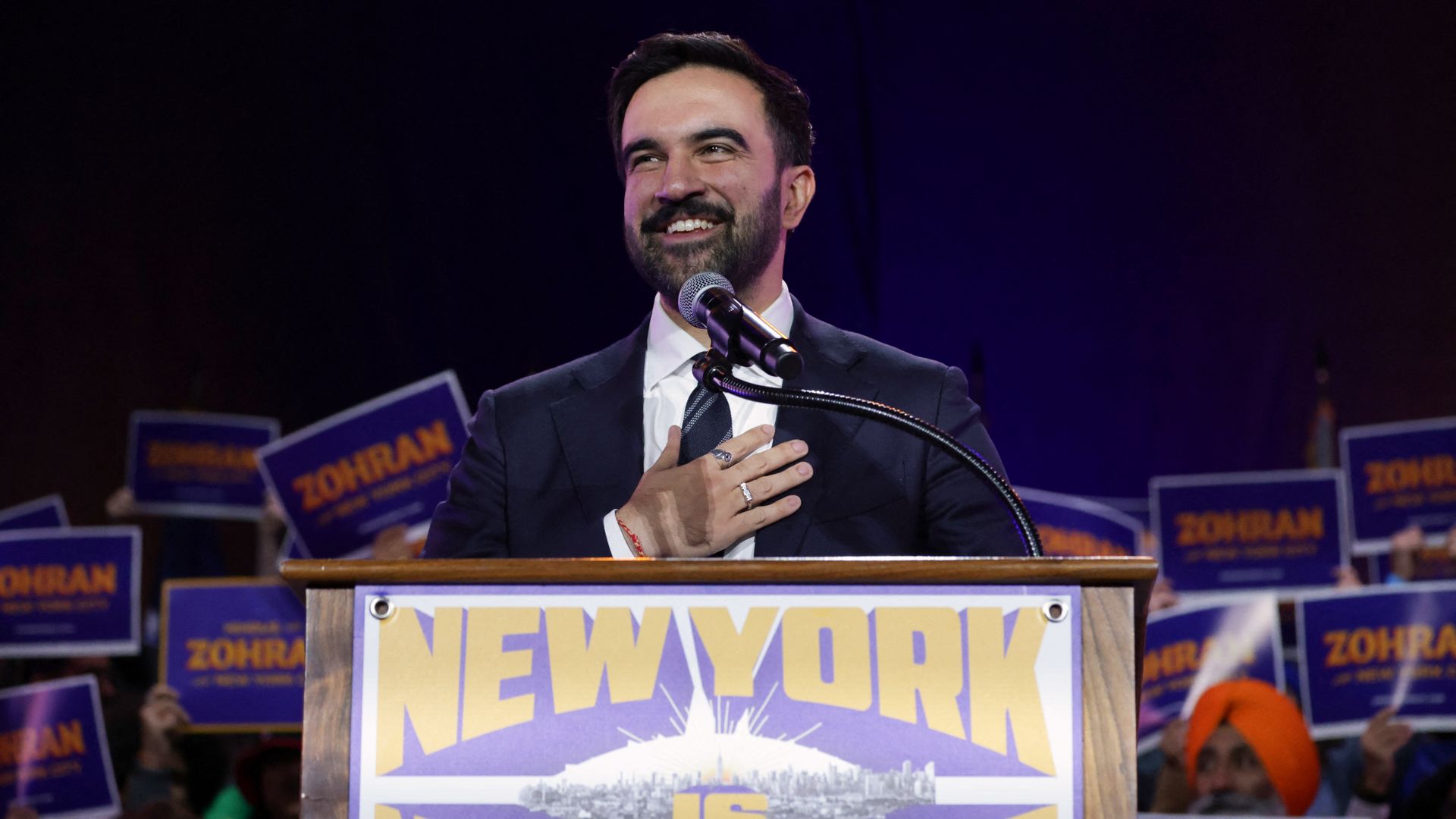 Zohran Mamdani smiling behind a podium that says "New York is Not for Sale," with people holding campaign signs behind him