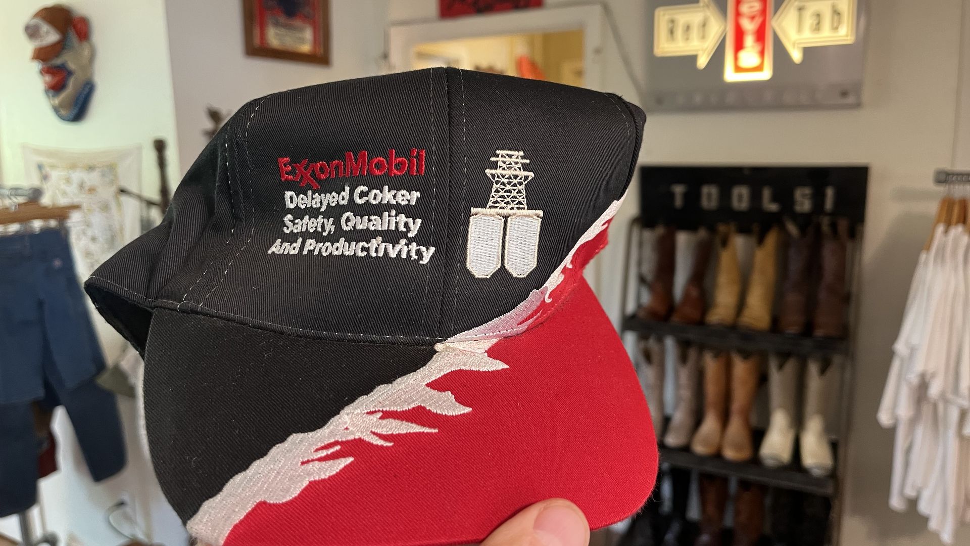 Close-up of a black cap with a red brim, embroidered with 'ExxonMobil' text, held by a hand in a clothing store; blurred background shows shoes and clothing on display.