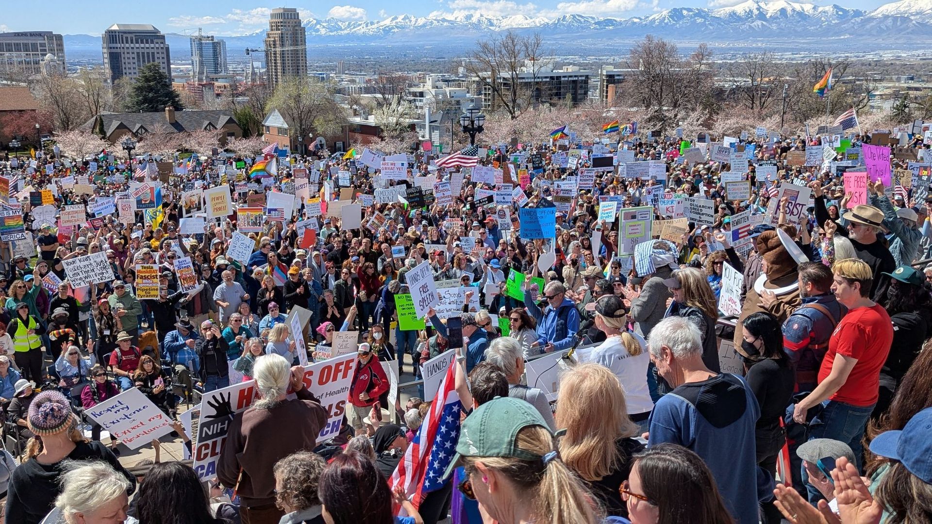 Thousands of protesters hold signs in a large outdoor space with city buildings and mountains in the background.